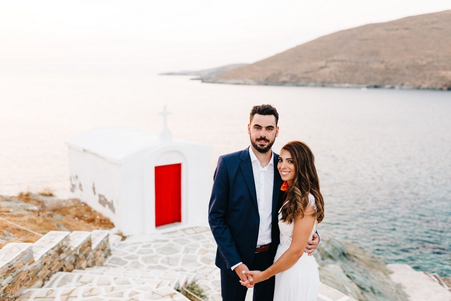 Newlyweds pose against white Mediterranean architecture and blue sea.