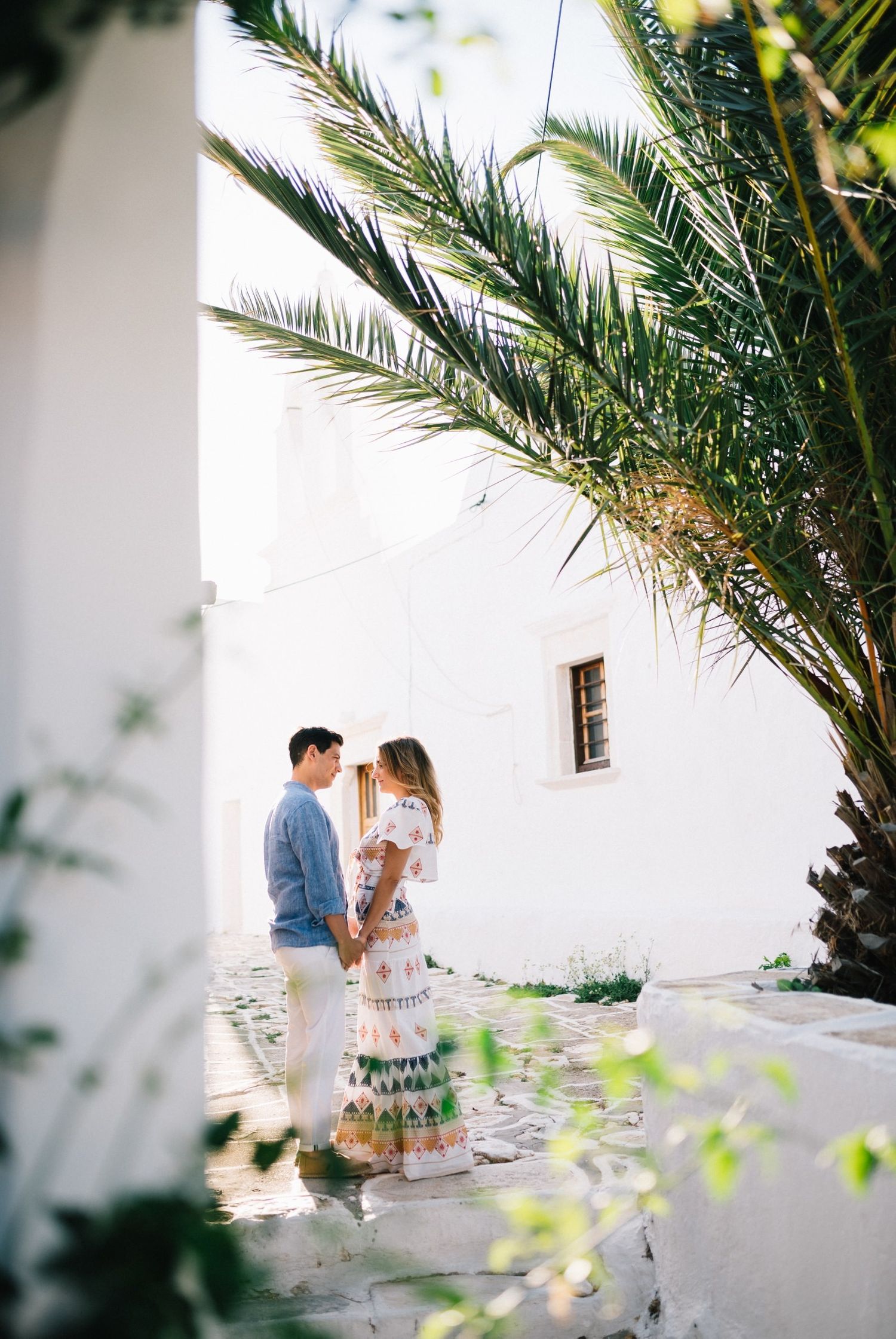 A romantic moment under palm fronds against a white Mediterranean building wall.