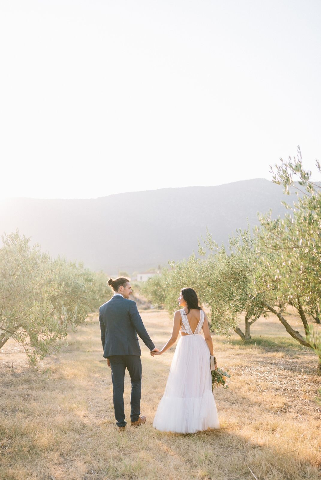 Newlyweds walking hand in hand through an olive grove with mountains in the background.