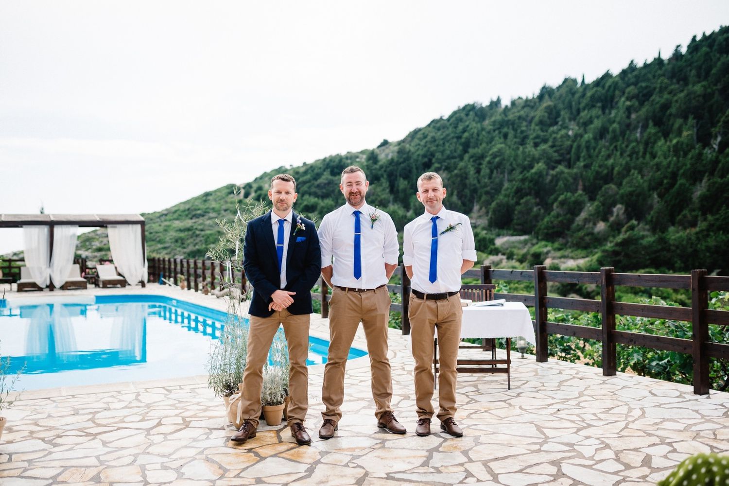 Three groomsmen in casual attire stand by a swimming pool with mountain views.