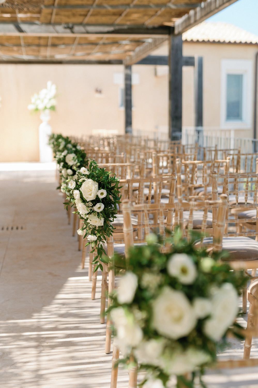 White flower arrangements line the aisle of an outdoor wedding ceremony with wooden chairs.