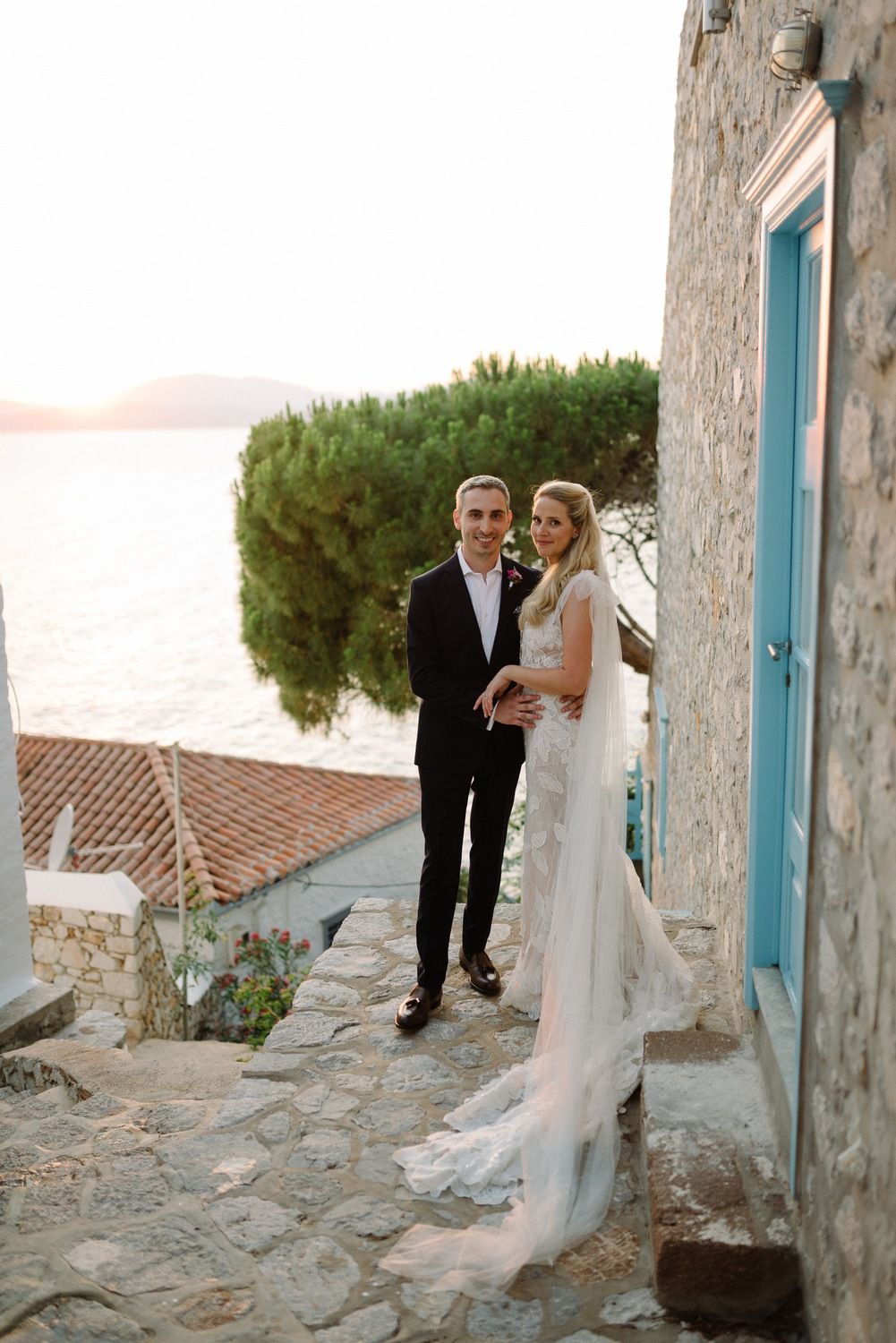 Wedding couple poses on charming stone path with Mediterranean cypress tree and sea view at sunset.