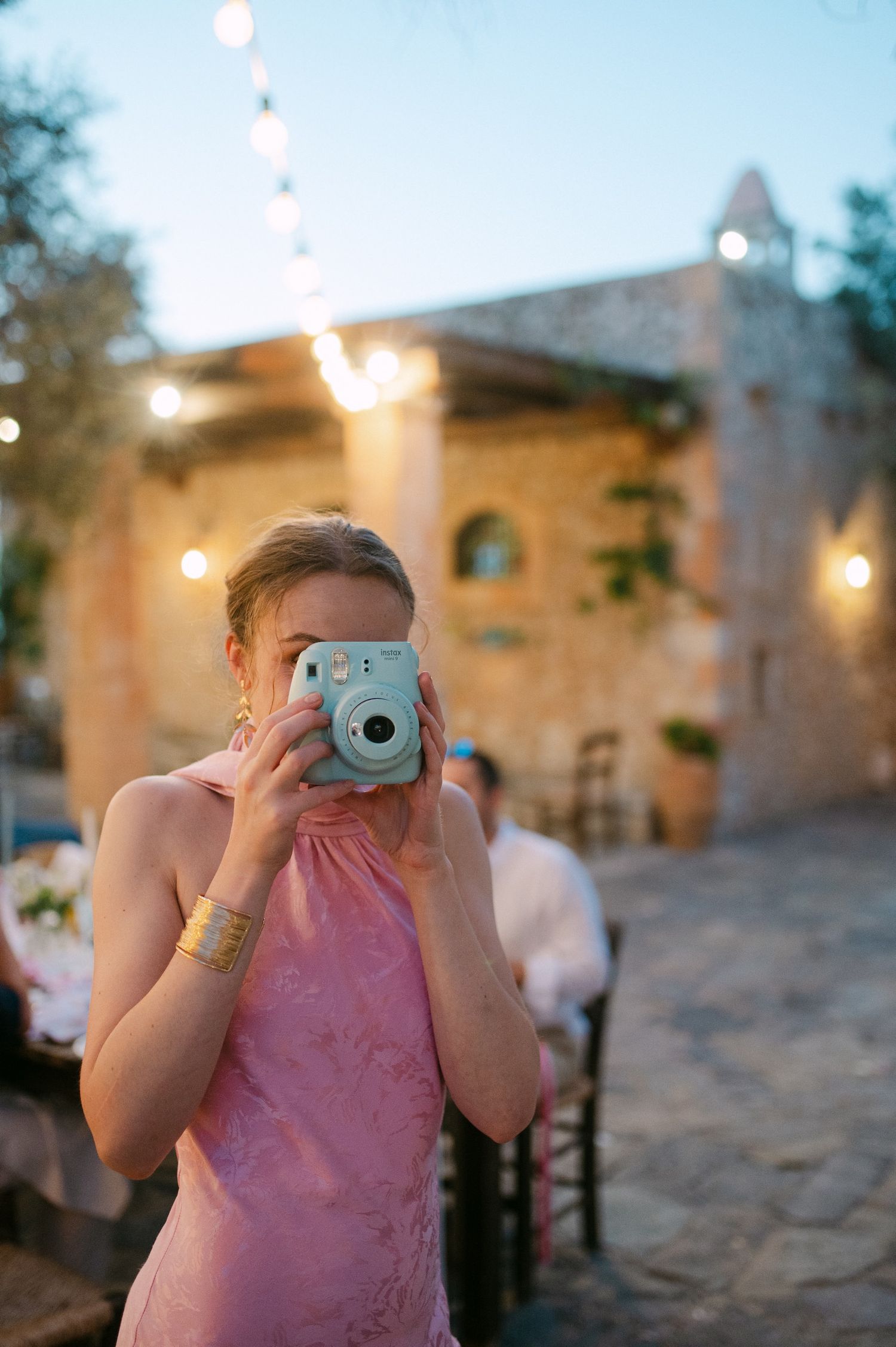 Someone takes a photo with a light blue Instax instant camera while wearing a pink dress at an outdoor evening event.