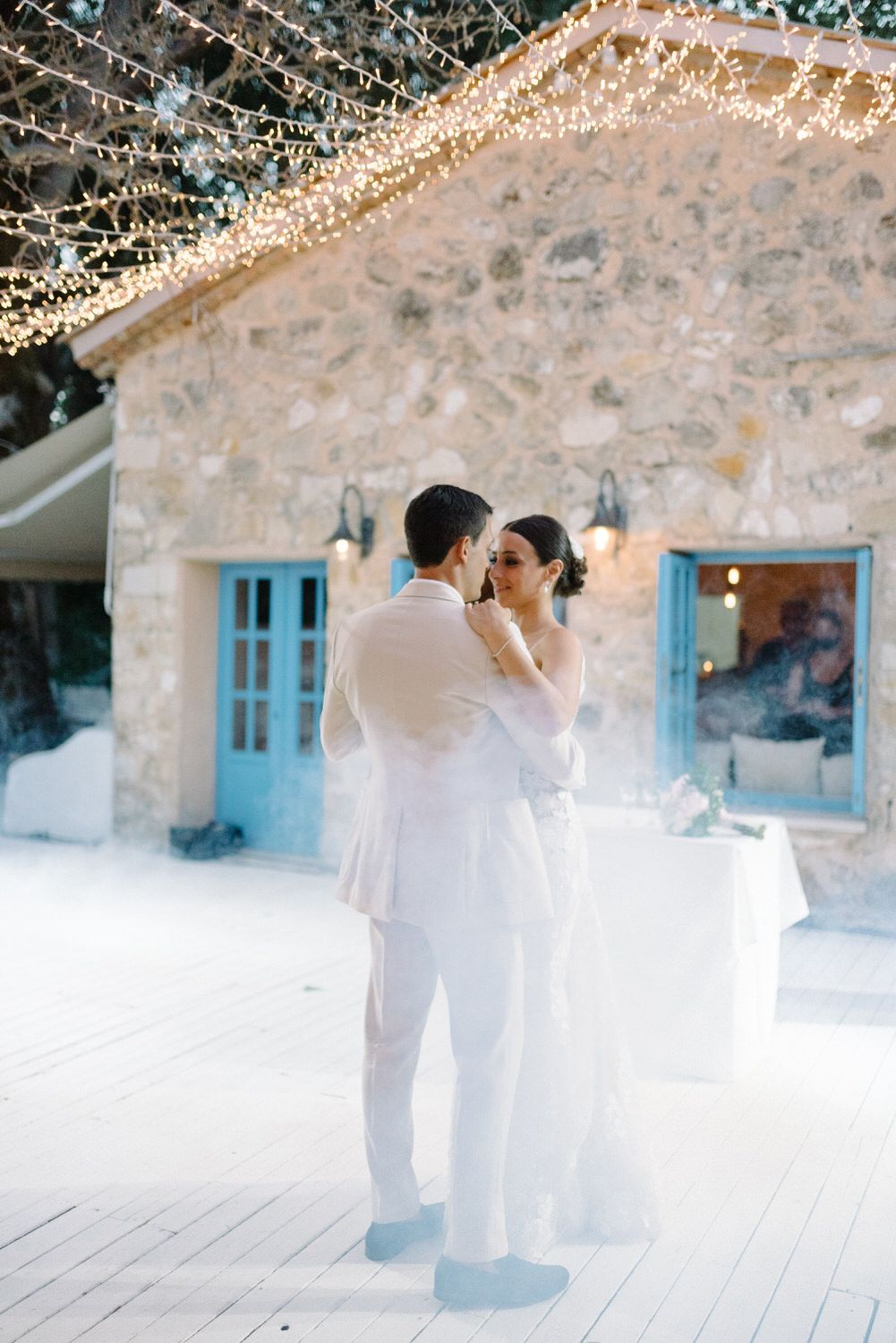 A couple dancing on a white deck under string lights with a rustic stone wall and blue door backdrop.