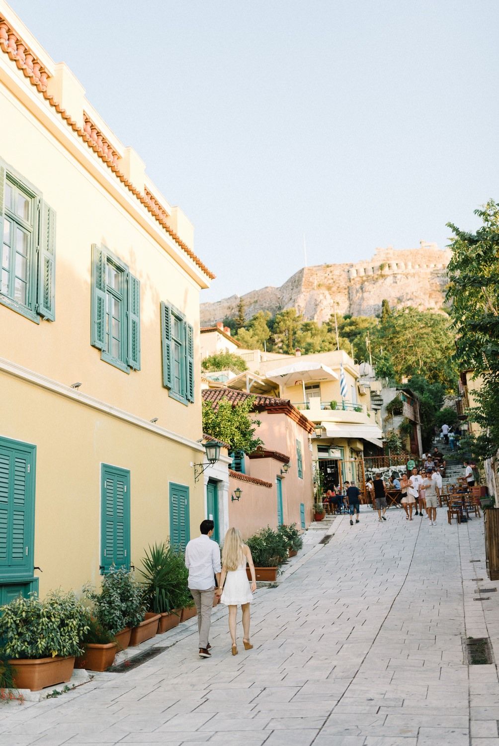 A couple walking down a charming Mediterranean street with pastel buildings and the Acropolis visible above.