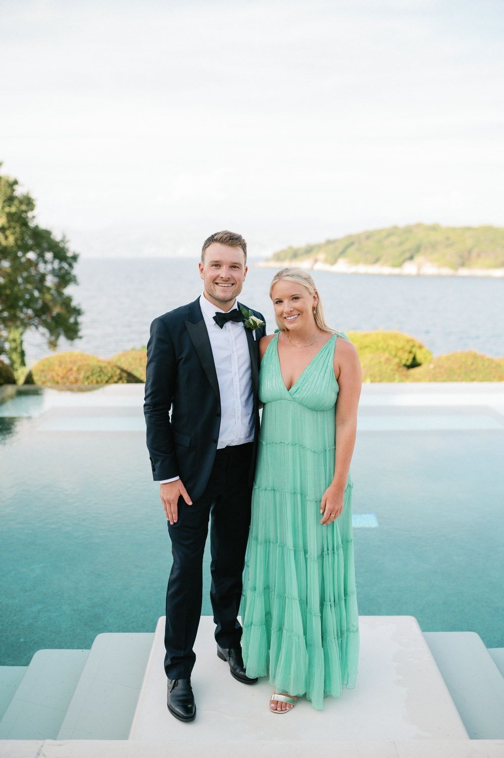 Couple poses by infinity pool overlooking ocean, wearing formal and mint green attire.