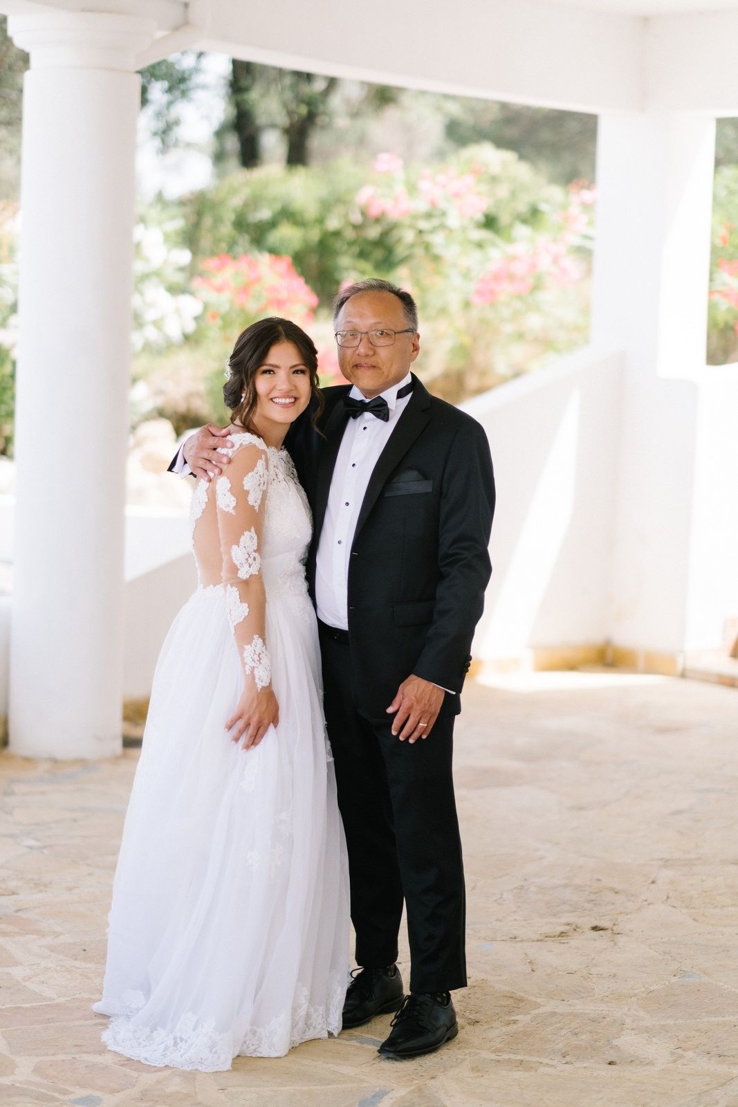 Elegant wedding portrait in white dress and tuxedo against garden backdrop.