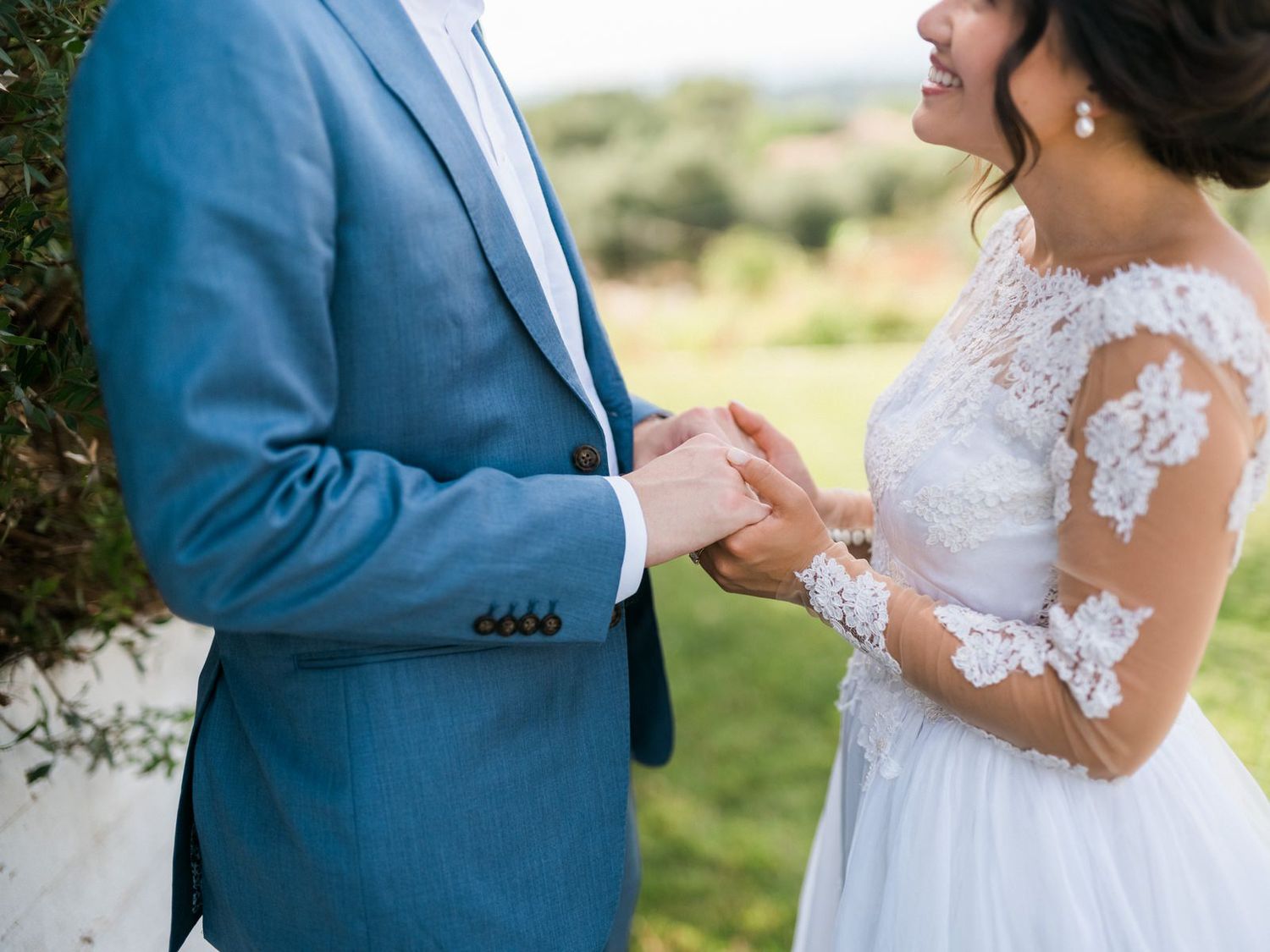 Close-up of couple holding hands outdoors with blue suit and lace wedding dress.