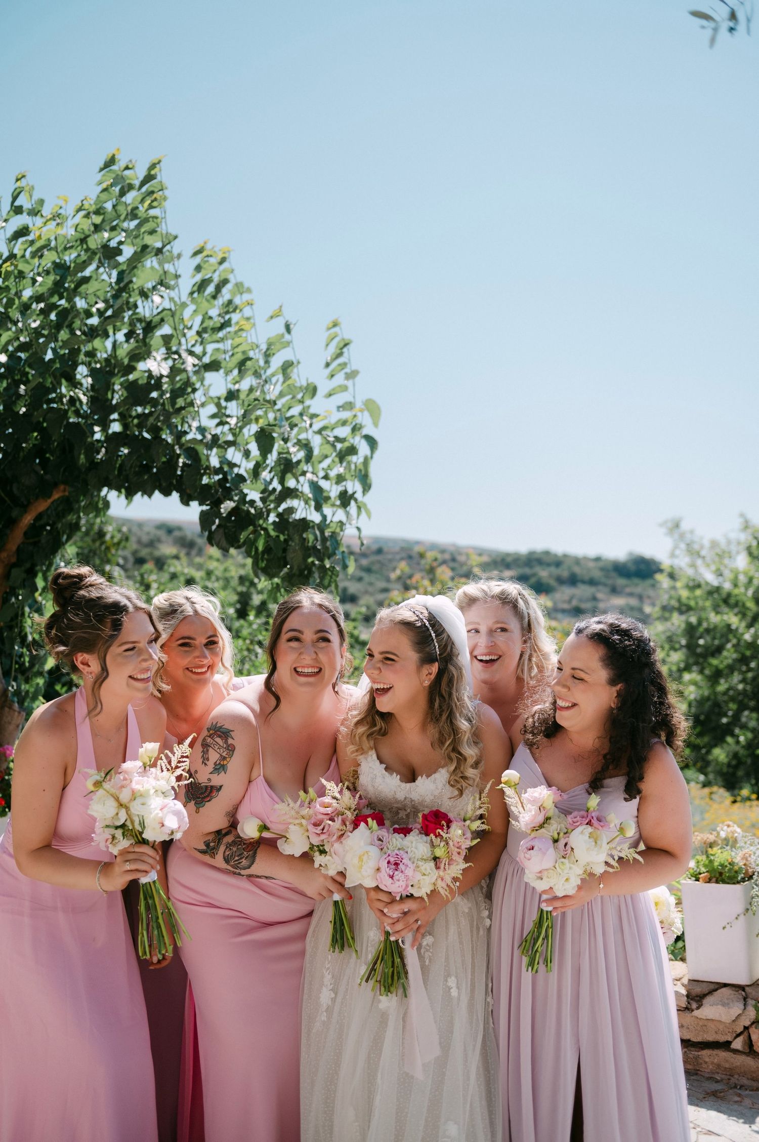 Bridesmaids in pink dresses laugh and share joyful moments while holding floral bouquets outdoors on sunny day.