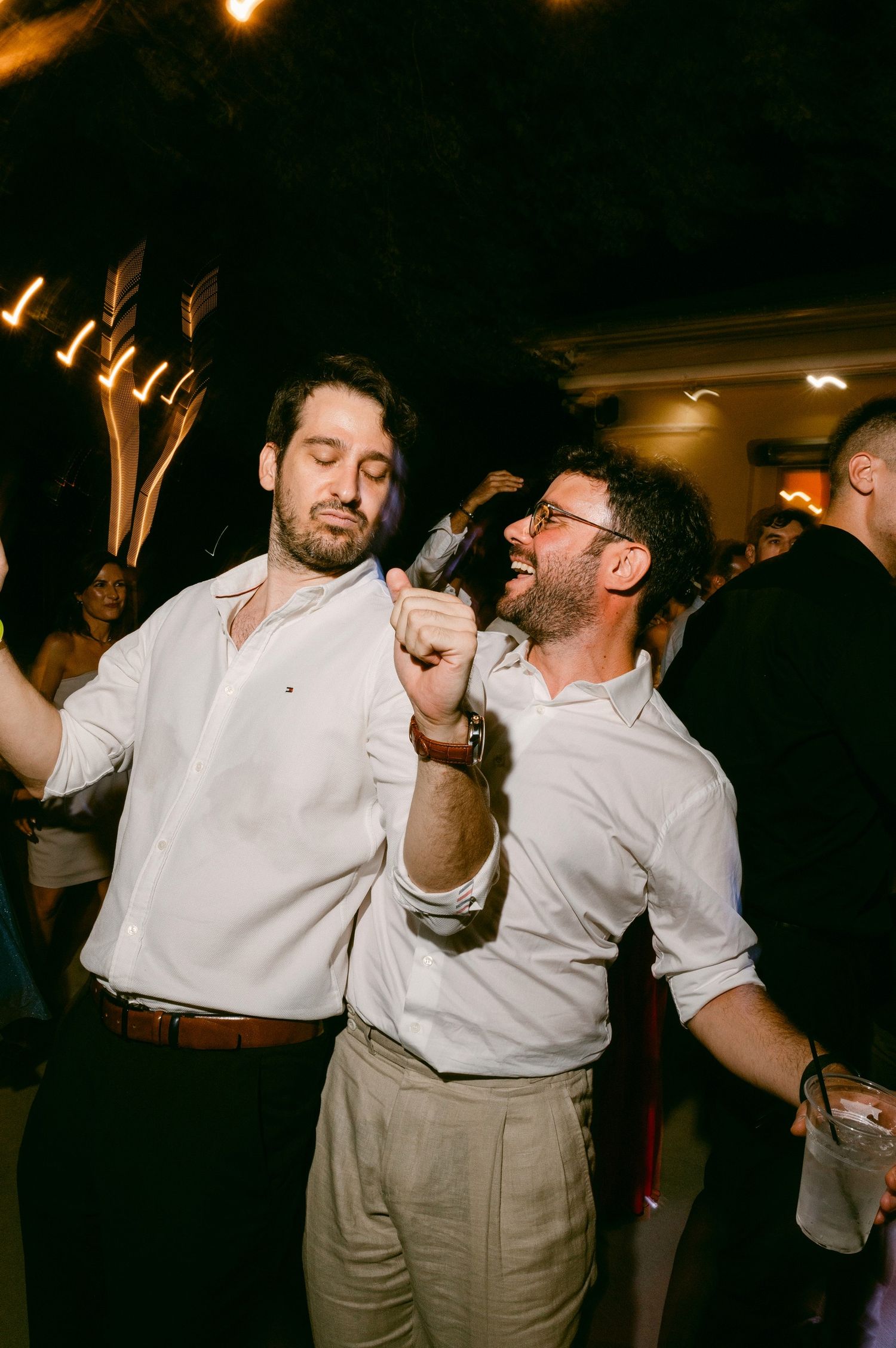 Two men in white button-down shirts dancing and singing enthusiastically at a party in a dimly lit room.