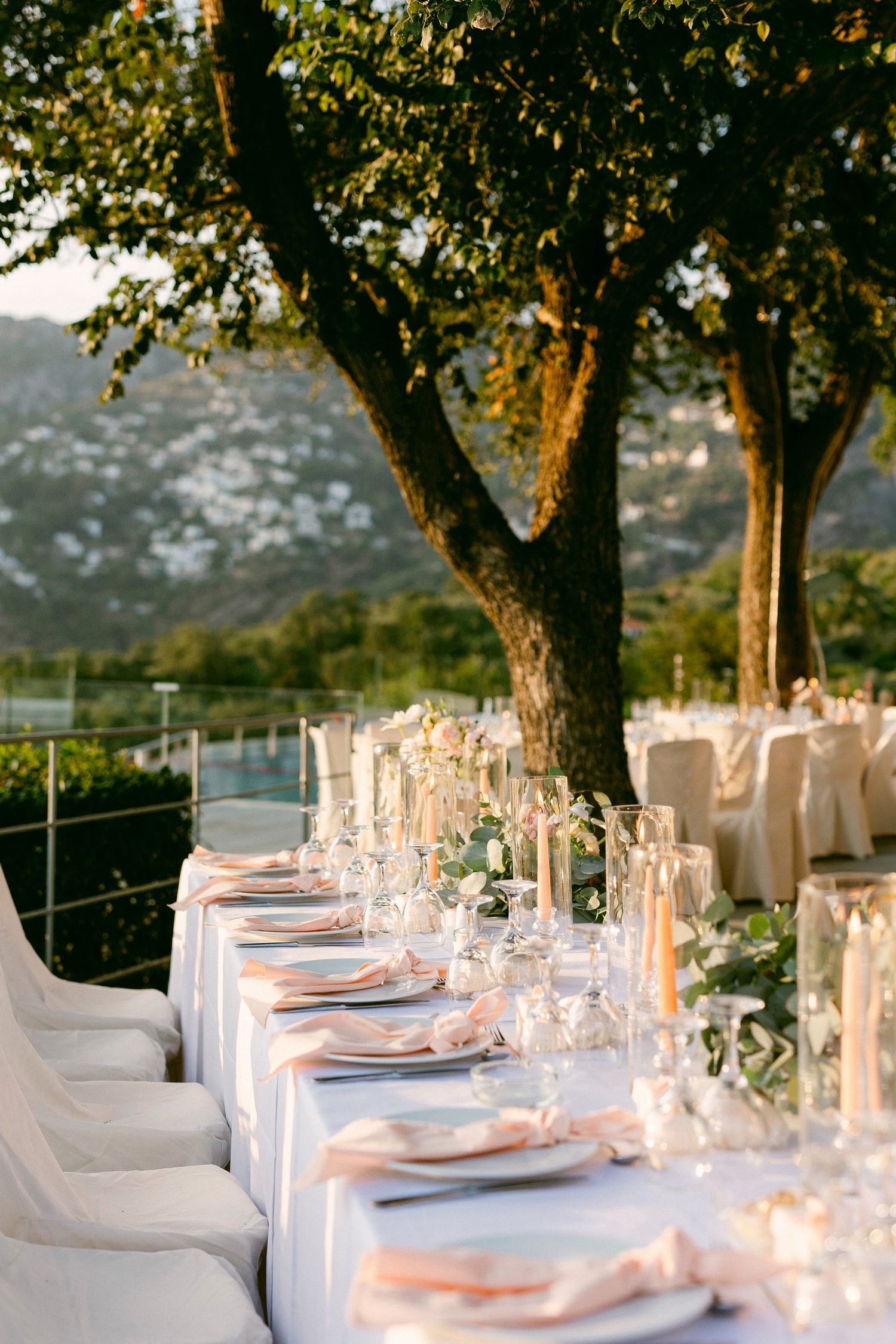 Elegant outdoor wedding reception table set with white linens and place settings under a tree at golden hour.