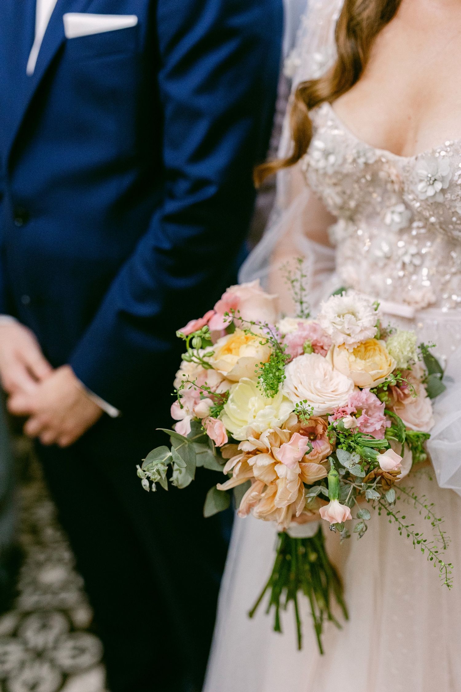 A delicate pastel bridal bouquet with pink and cream blooms held against a navy suit and white wedding dress background.