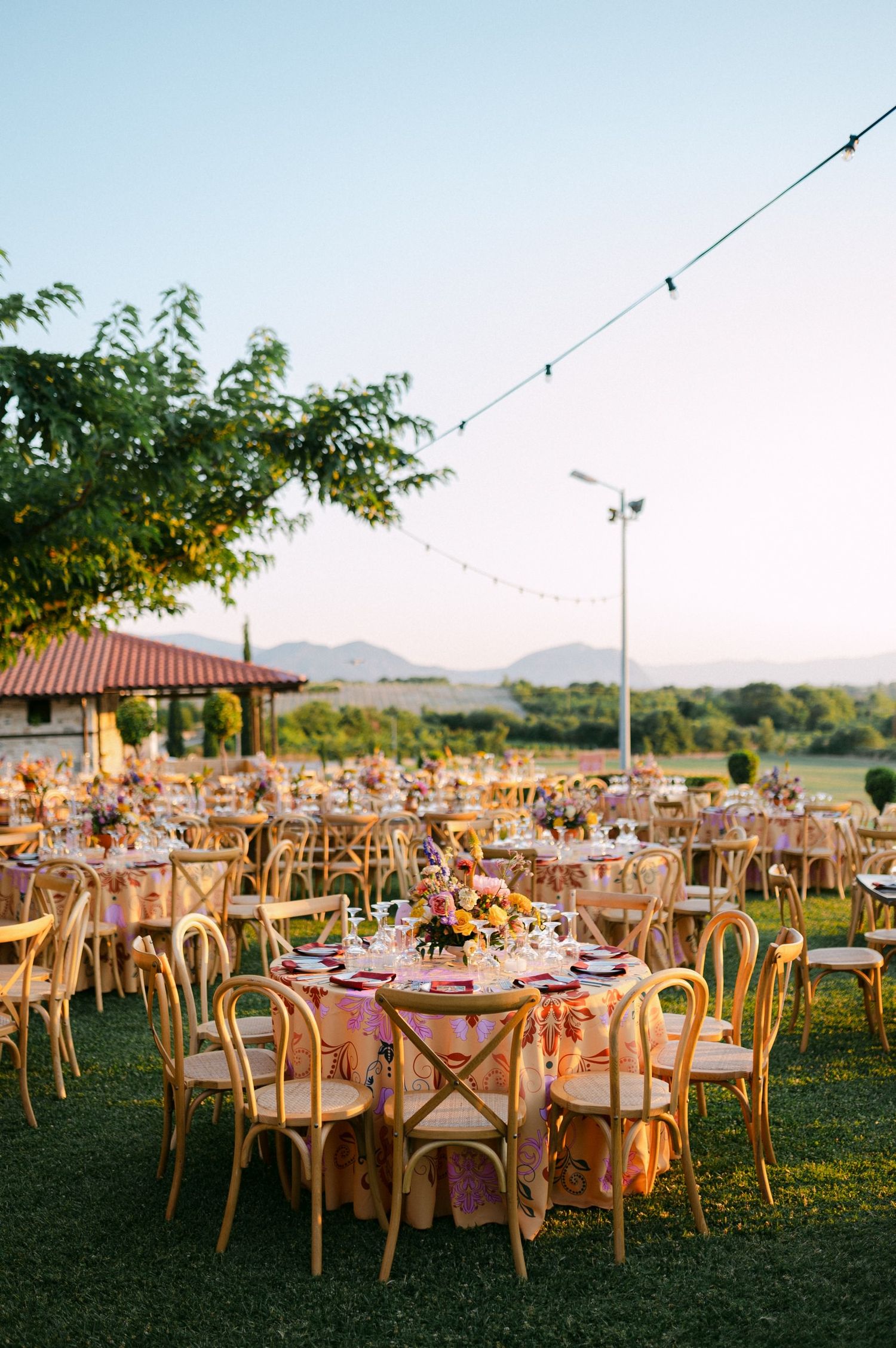 Elegant outdoor wedding reception setup with round tables and wooden chairs on a grassy lawn at sunset in vineyard Kamkoutis Velvento Greece