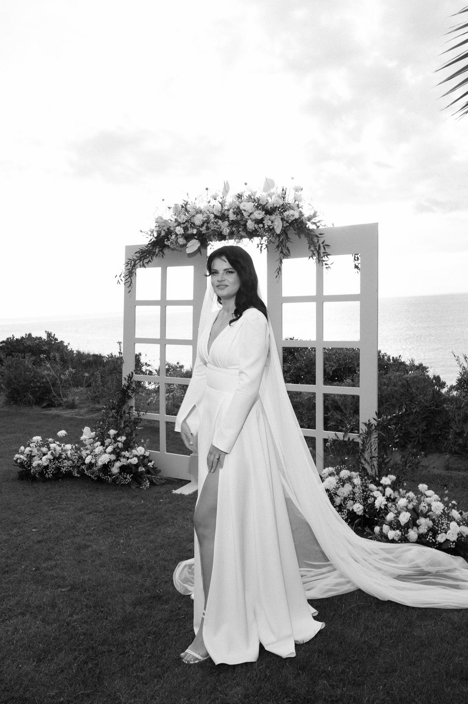 A bride in a modern white wedding jumpsuit with flowing cape stands by a floral arch overlooking the ocean.