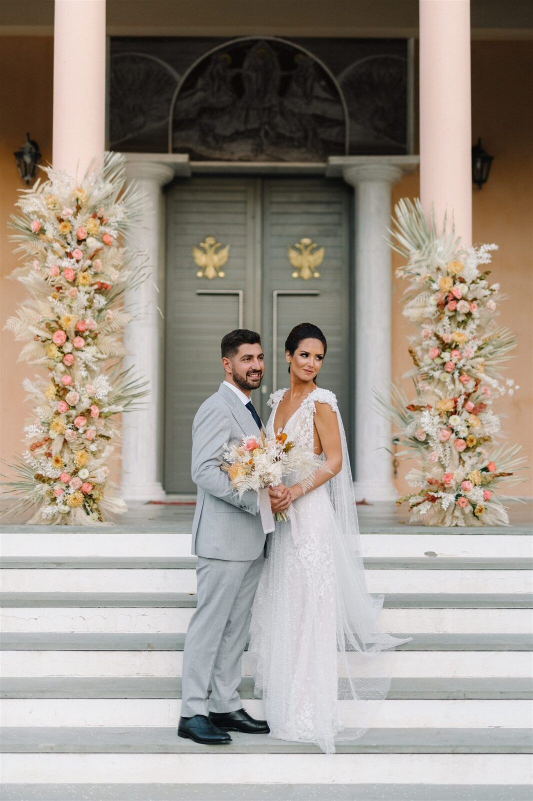 A romantic wedding couple poses on white steps with elegant peach and white floral arrangements decorating the columns.