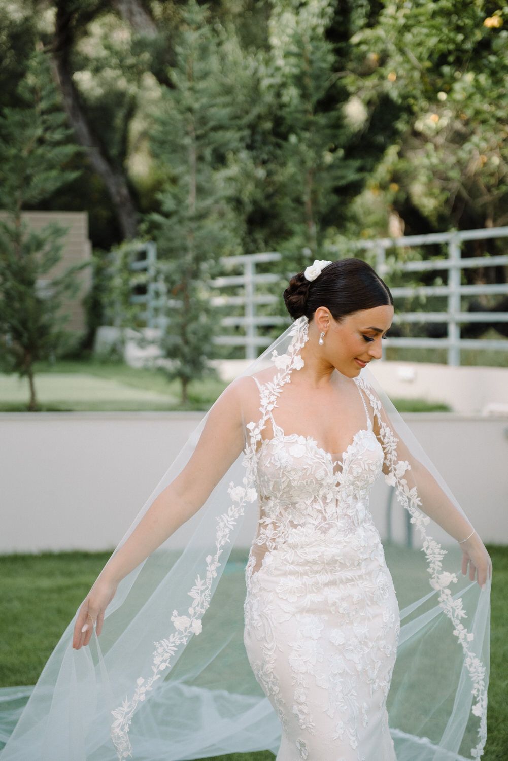 Bride wearing stunning lace wedding dress adjusts flowing veil in outdoor garden.