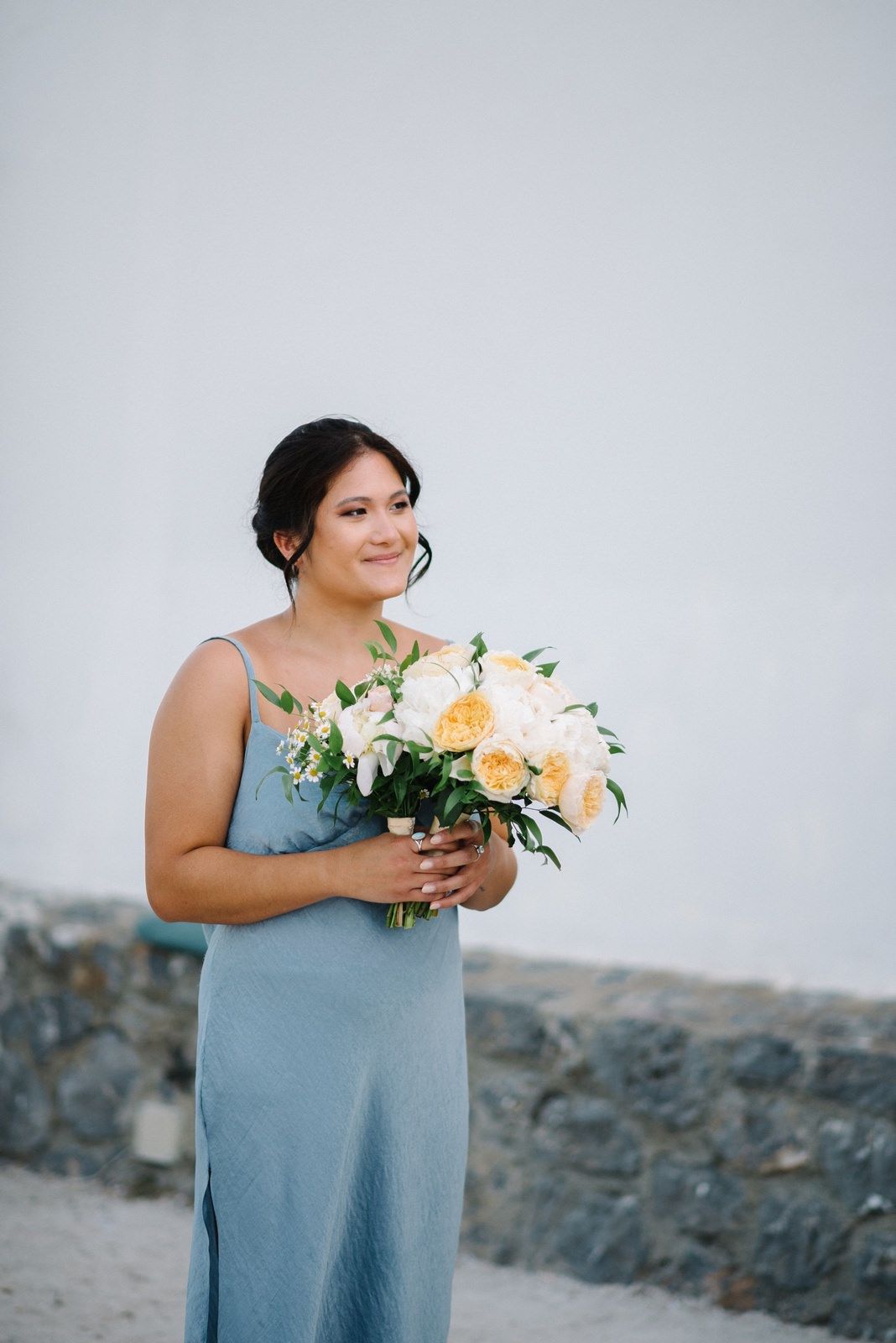 Woman in blue dress holding peach and white floral bouquet against stone wall.