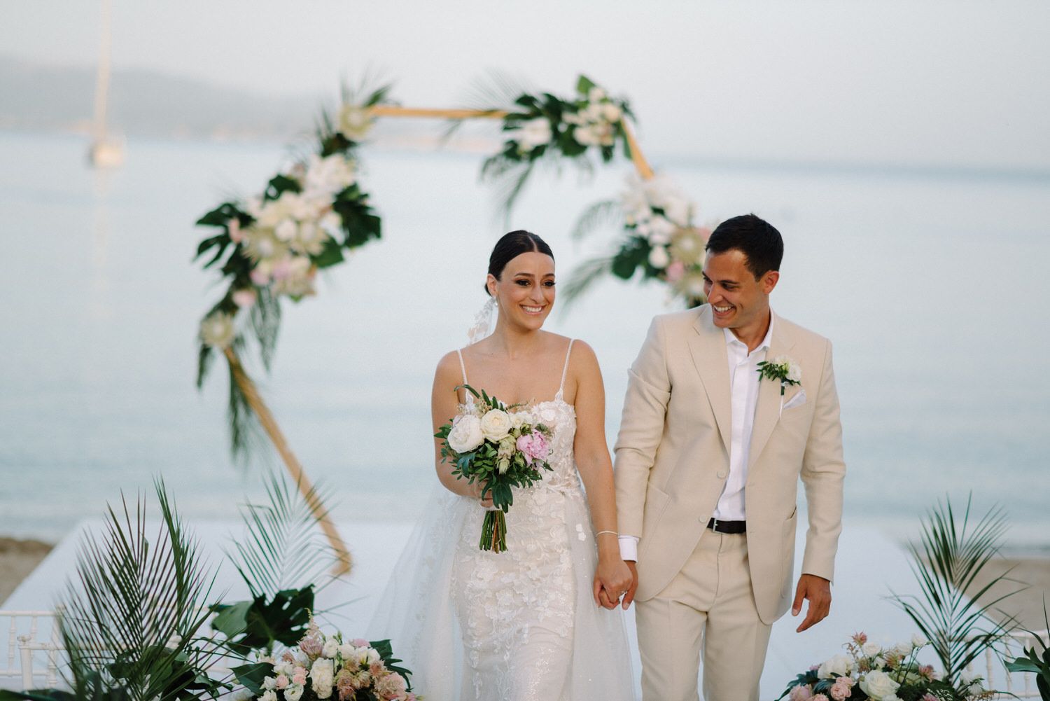 Newlyweds holding hands under hexagonal arch with white flowers and ocean view.