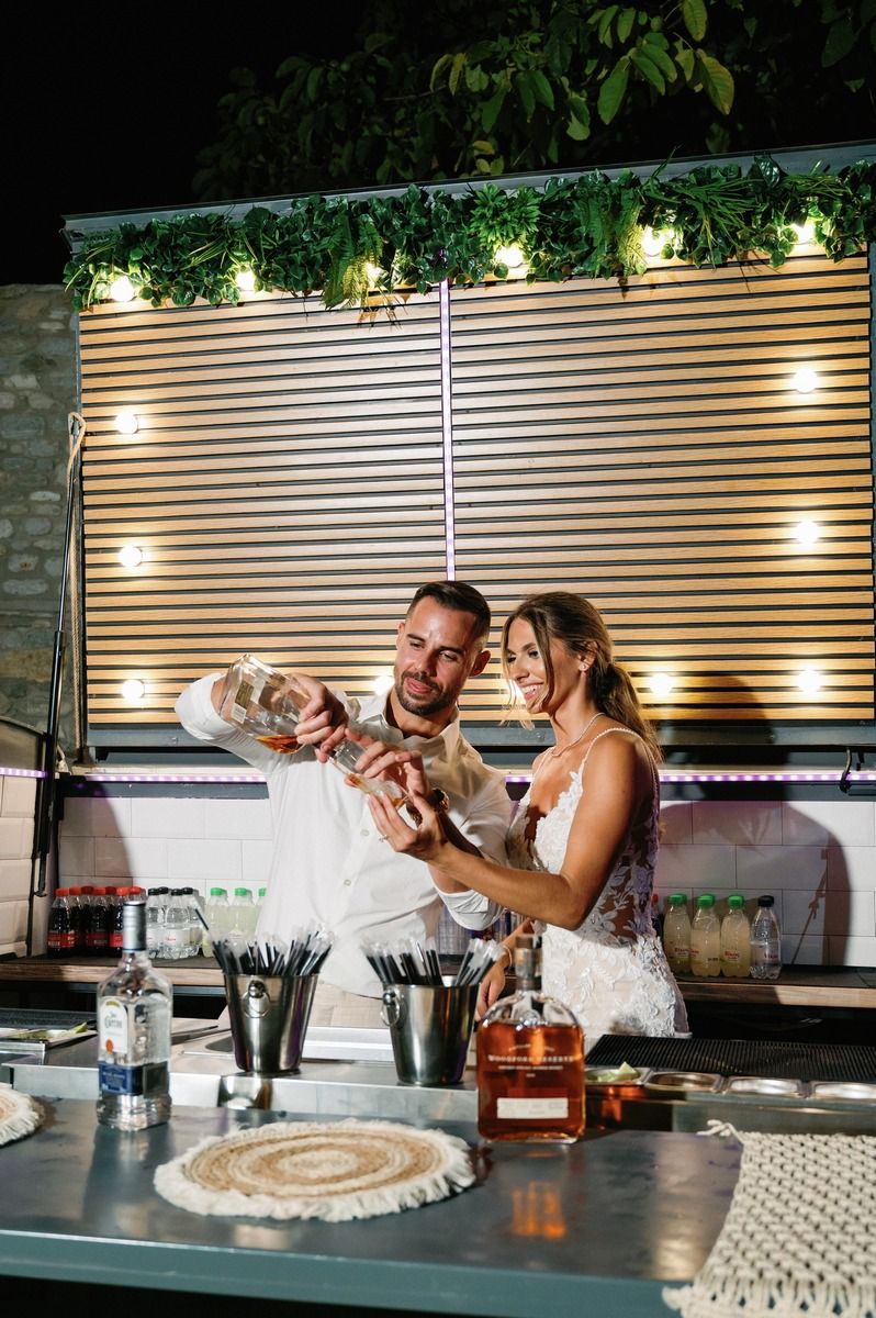 Bartenders prepare cocktails together at an outdoor bar with wooden slat backdrop and ambient lighting.