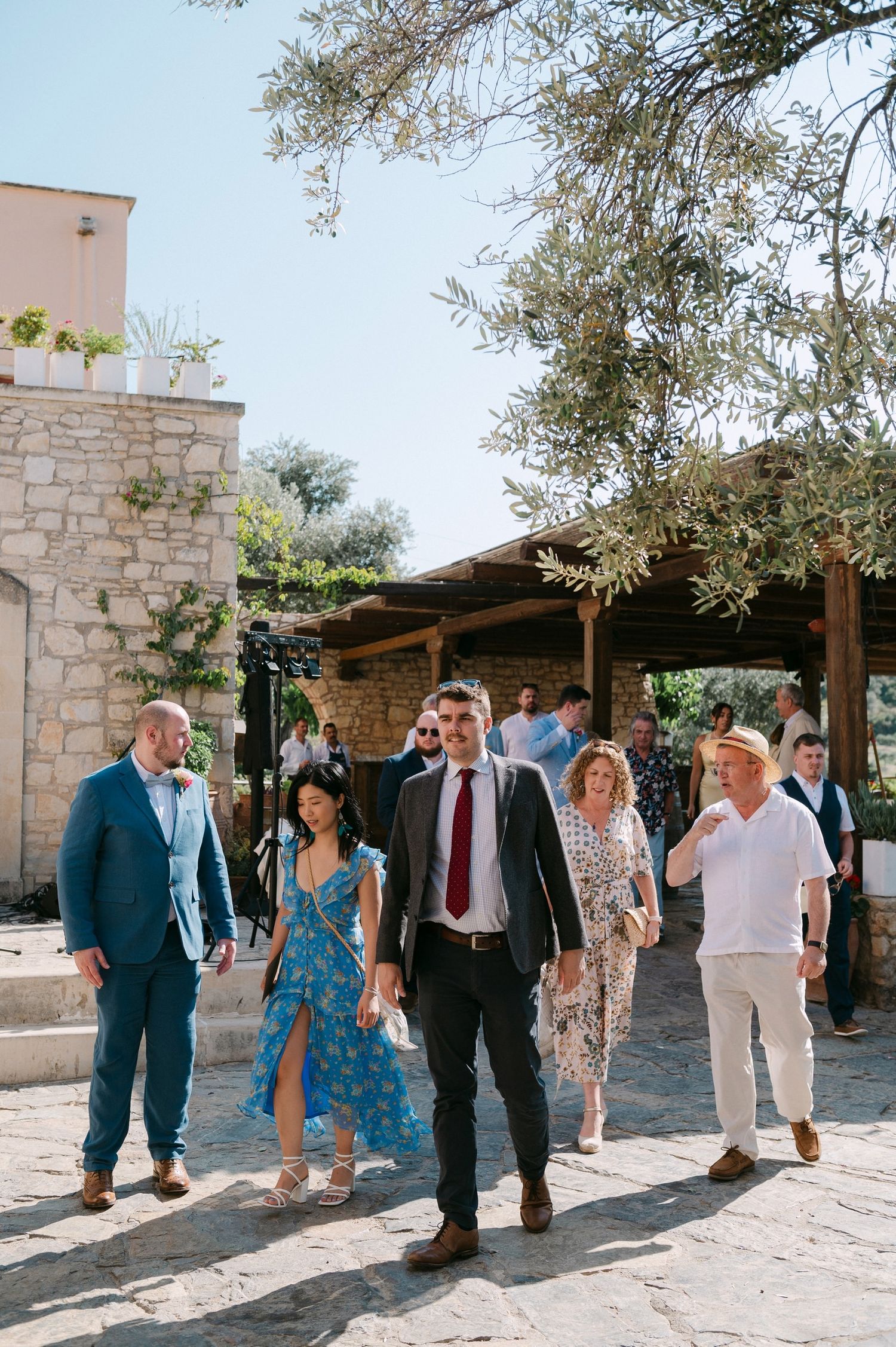 Wedding guests walk and socialize under an outdoor pergola with stone walls and olive trees.