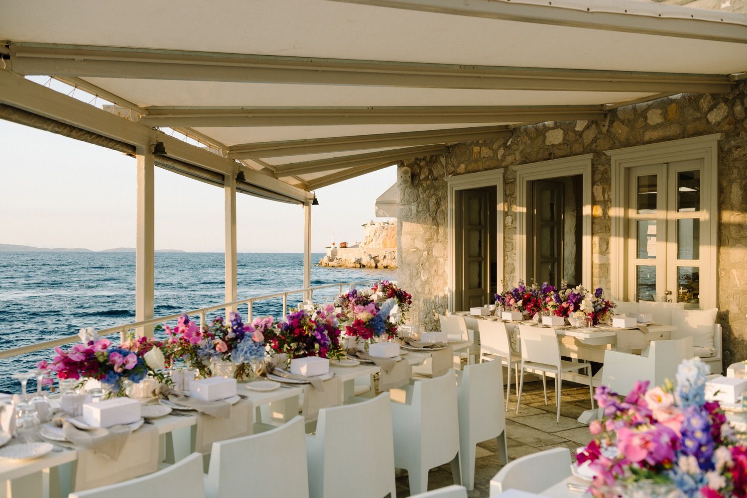Elegant waterfront dining setup with long table decorated in pink and purple floral arrangements overlooking the ocean.
