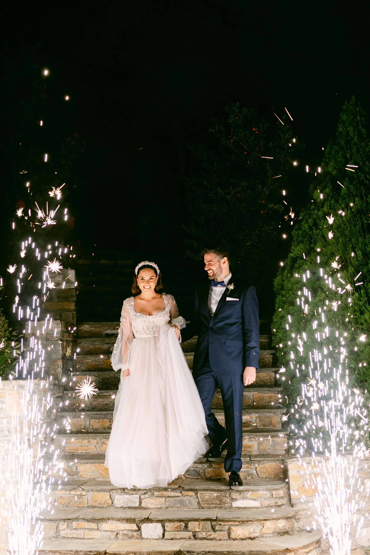 A couple descends stone stairs at night with glowing sparkler fountains creating magical light effects on both sides.