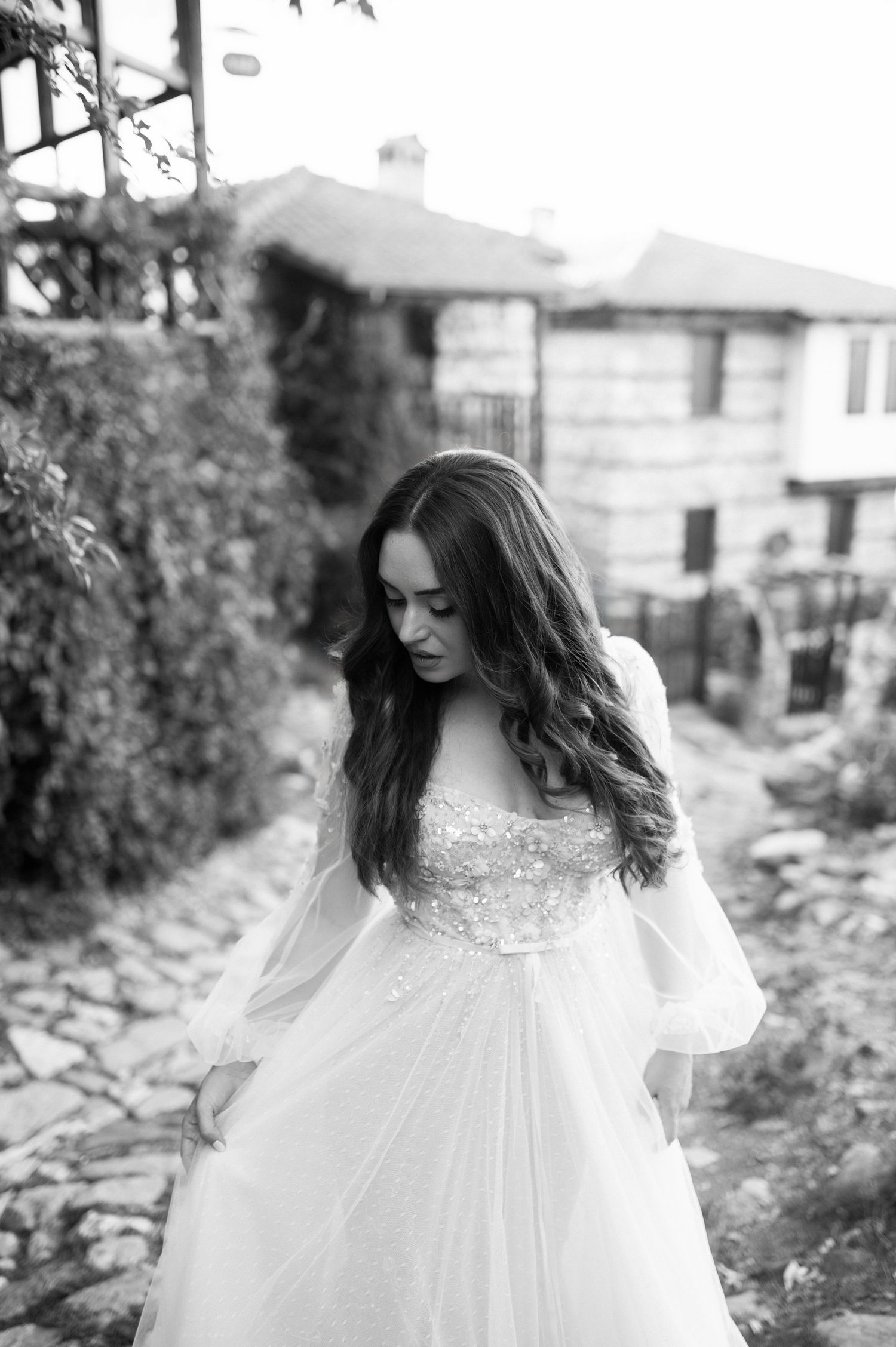 A person in a flowing white wedding dress with long sleeves stands on a cobblestone path beside a garden.