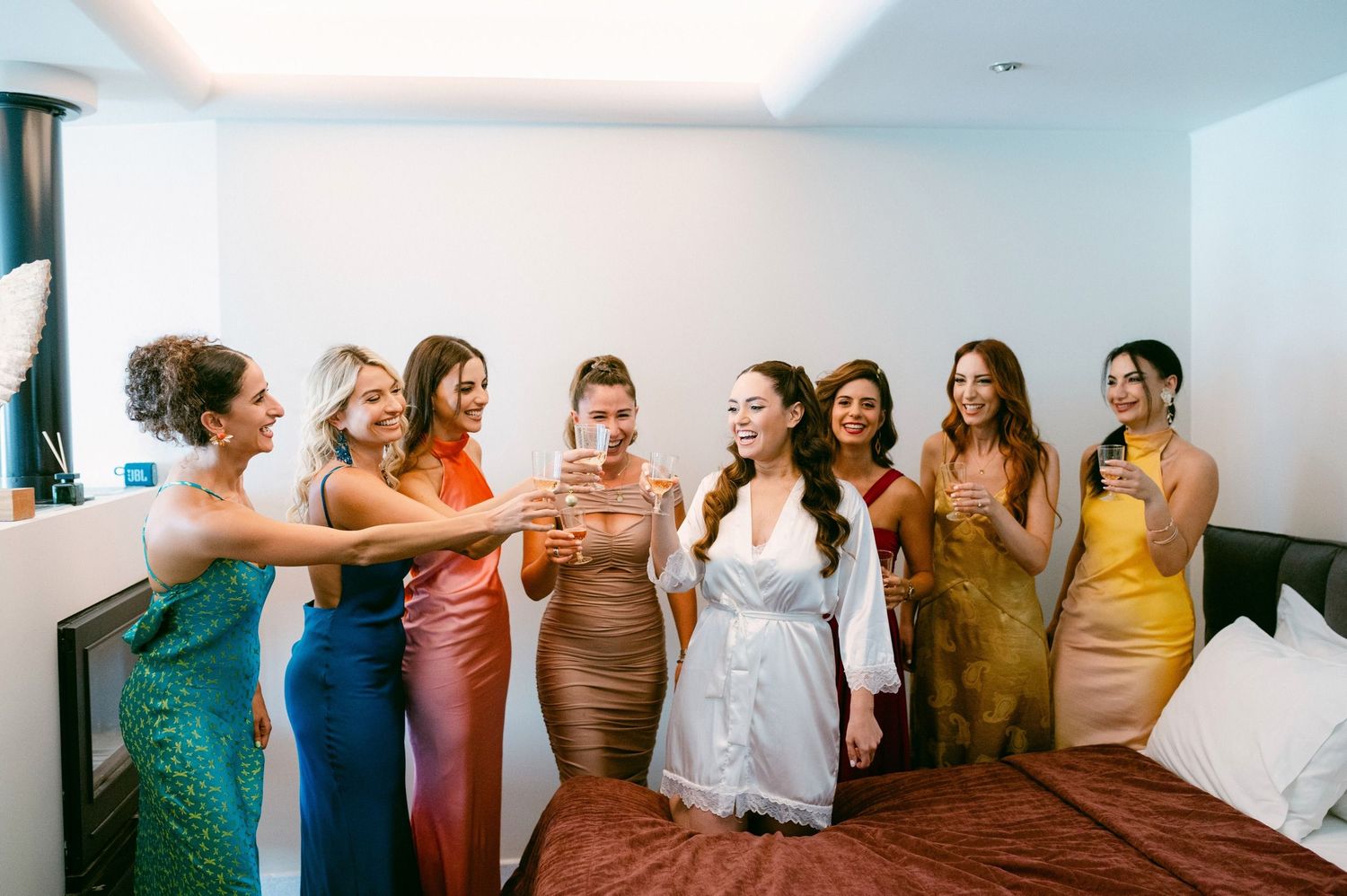 A group of women in colorful formal dresses toast with champagne glasses in a bright bedroom setting.