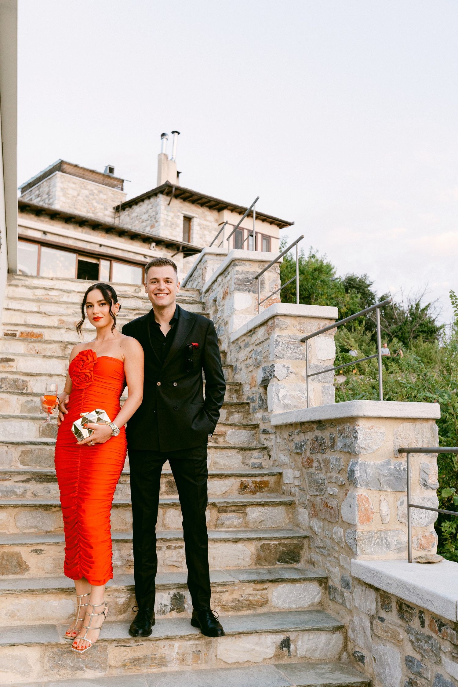 A couple poses on stone steps outside a Mediterranean-style villa, wearing formal red and black attire.