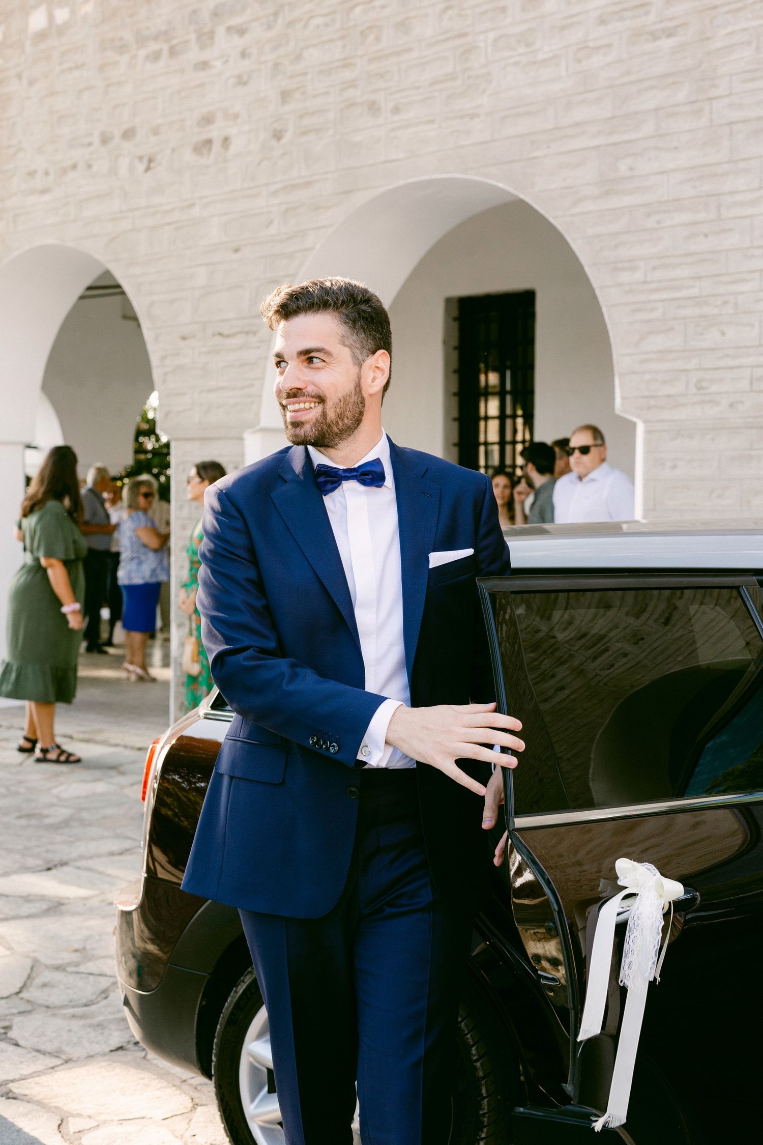 Groom in navy blue suit and bowtie stands next to classic car outside arched building during wedding celebration.