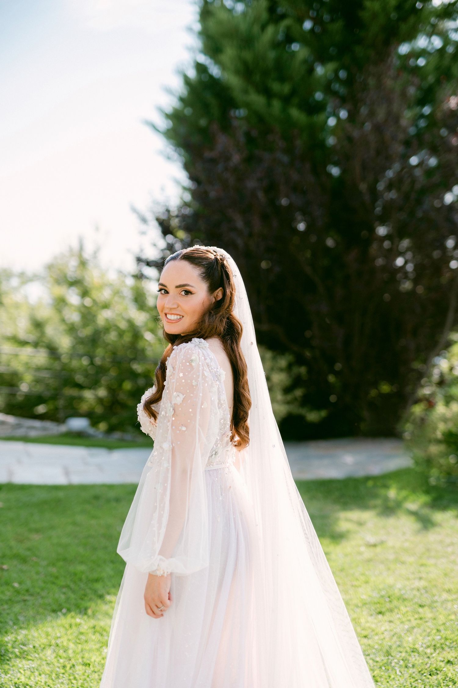 A bride in a white long-sleeved wedding gown with lace details and flowing veil poses outdoors on a sunny day.