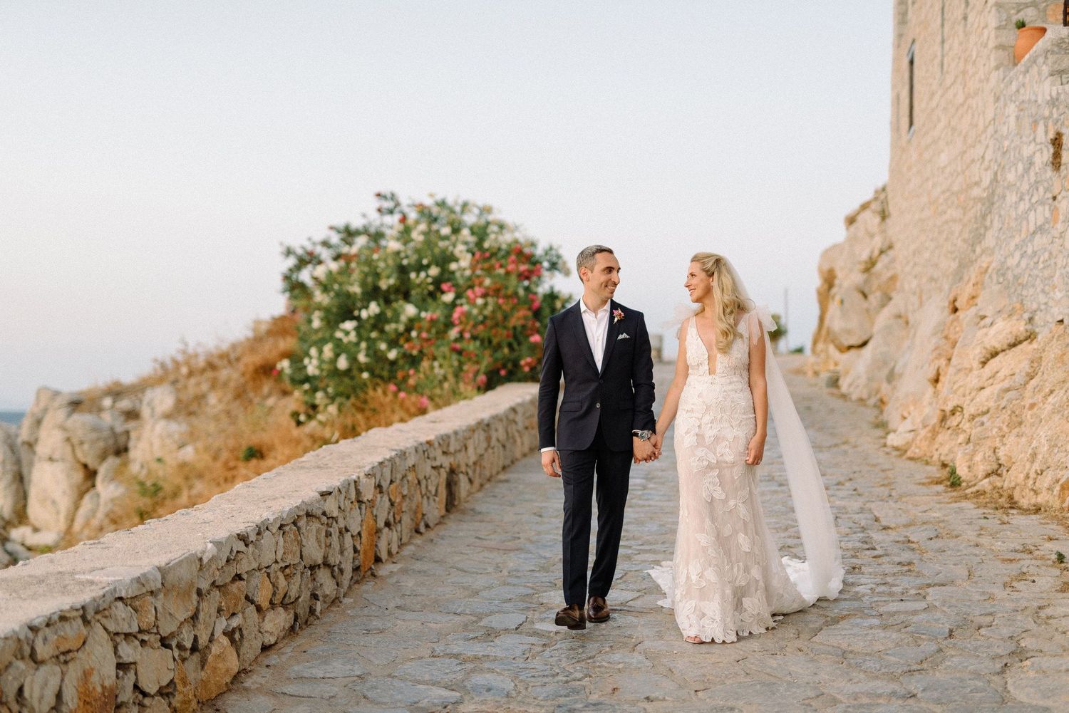 A couple walks along a rustic stone pathway in a Mediterranean destination during their wedding photoshoot.