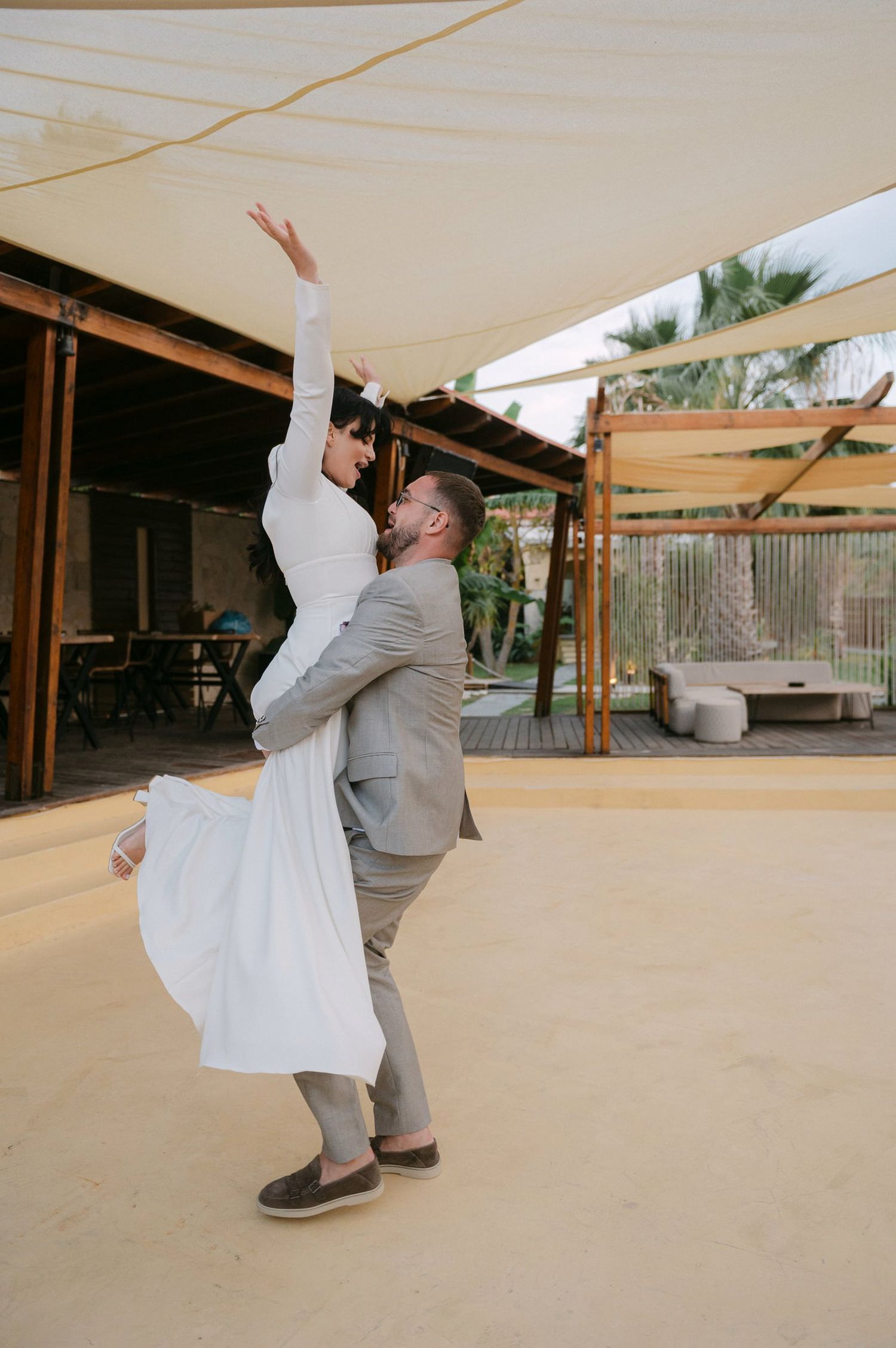 A couple performs a graceful dance move outdoors under a covered patio with palm trees visible in the background.