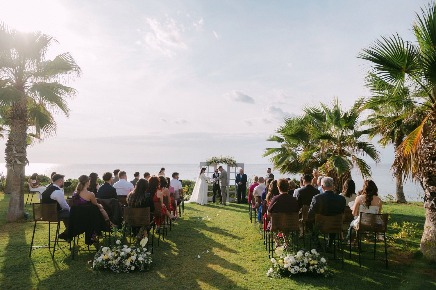 Panoramic view of an outdoor wedding ceremony setup with palm trees and guests seated on a lush green lawn at sunset.