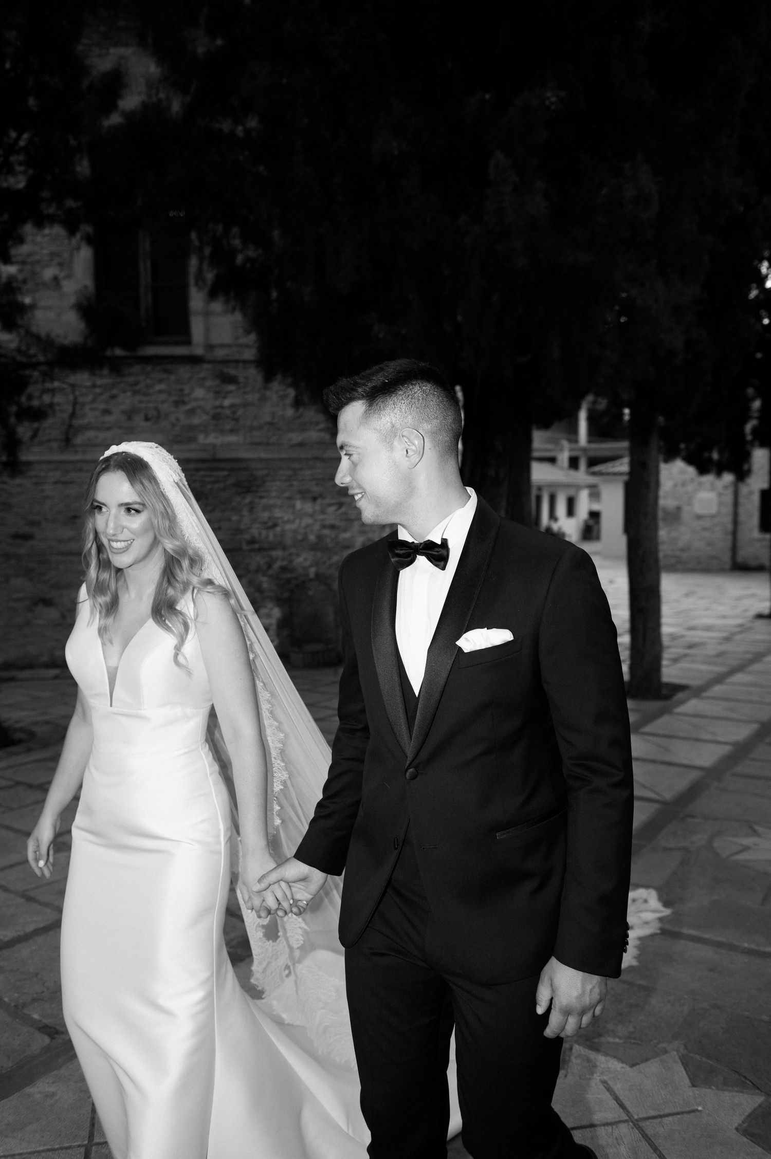 A black and white wedding photograph of a bride in a sleek white dress and groom in tuxedo walking together outdoors.