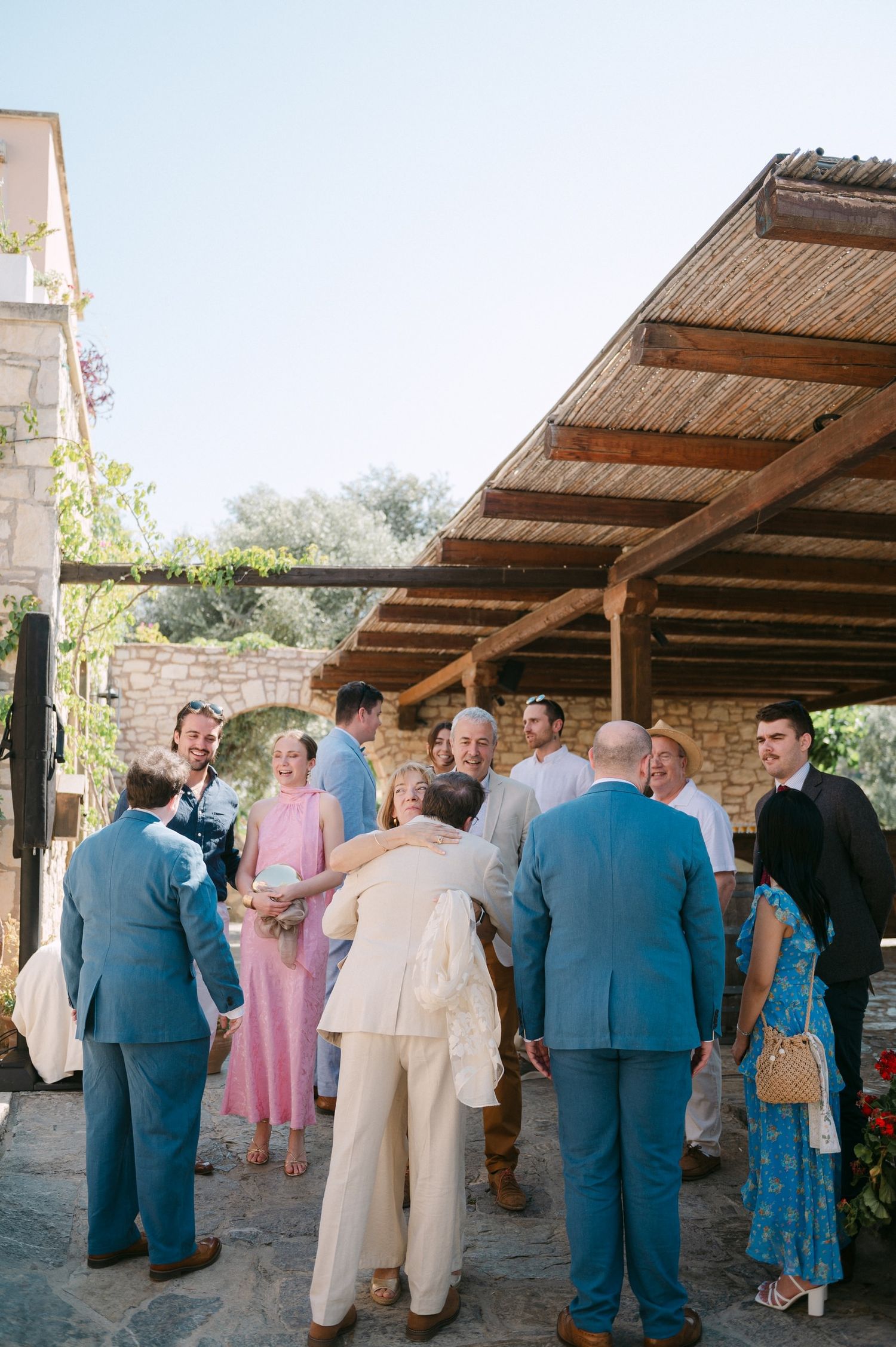 Guests mingling and socializing under a rustic wooden pergola at an outdoor wedding reception.