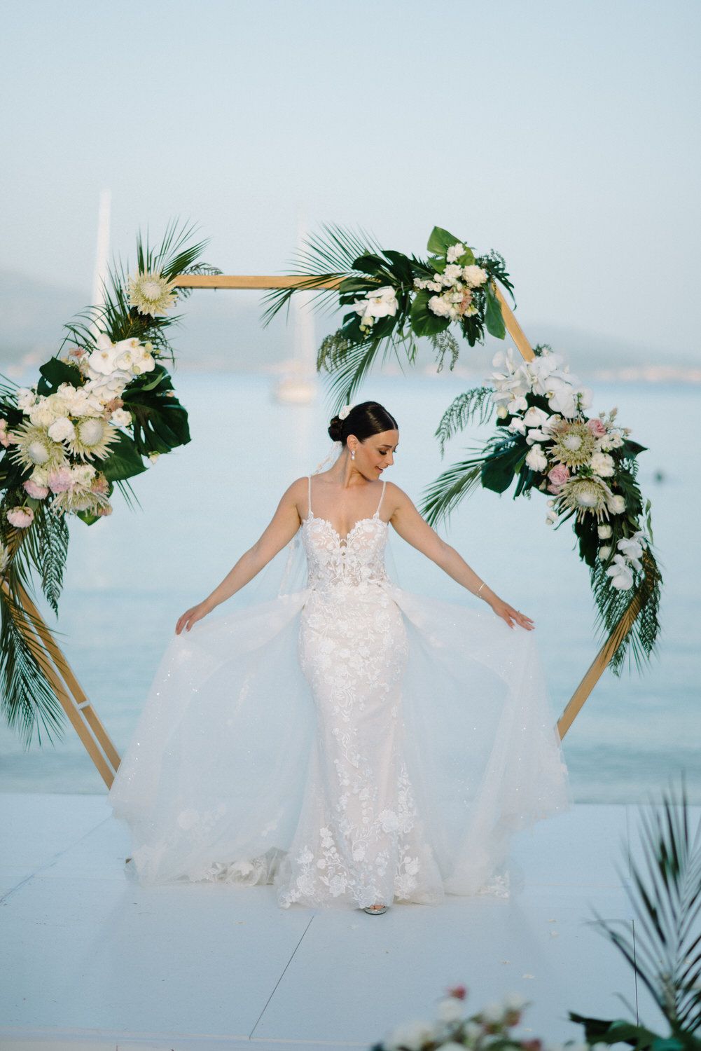 Bride in lace wedding gown poses under floral hexagon arch with ocean view.