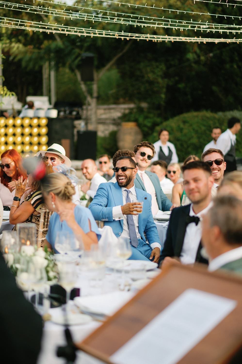 Wedding guests enjoy an outdoor reception under string lights with drinks and laughter at elegant table settings.