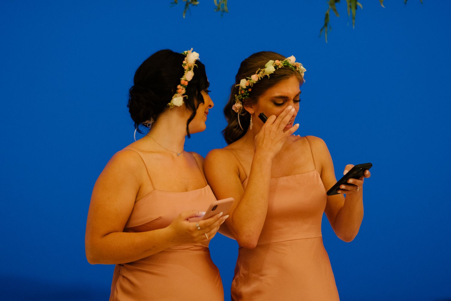 Two attendants in coral dresses look at their phones against a deep blue evening sky.
