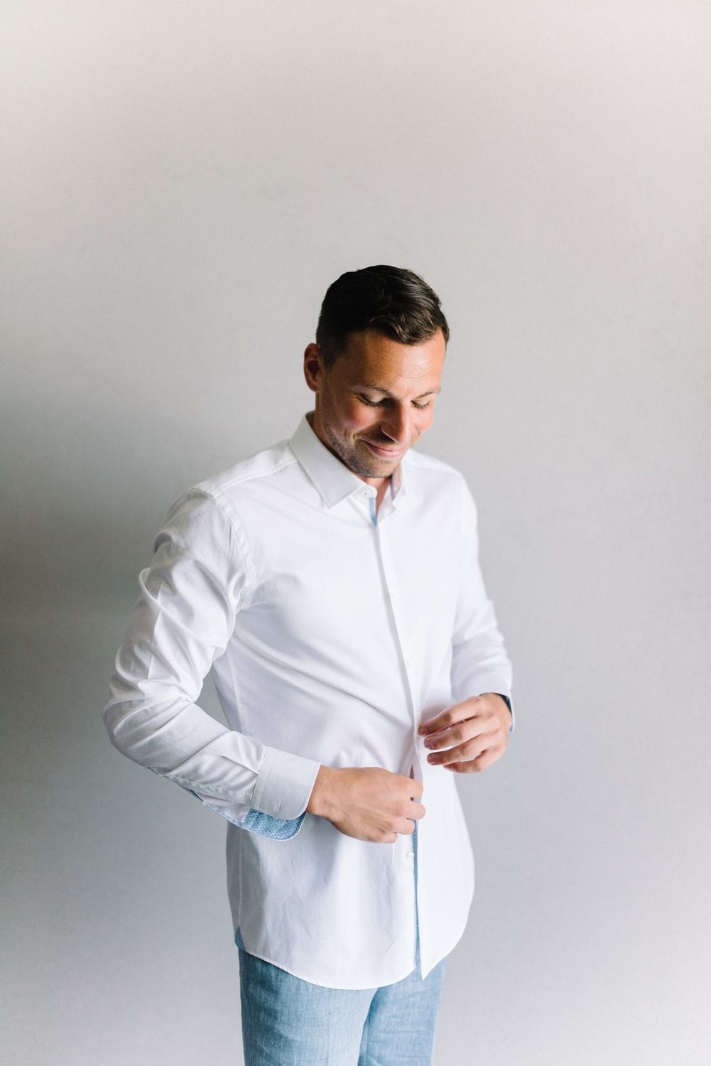 Man in crisp white button-down shirt buttoning his sleeve against neutral background.