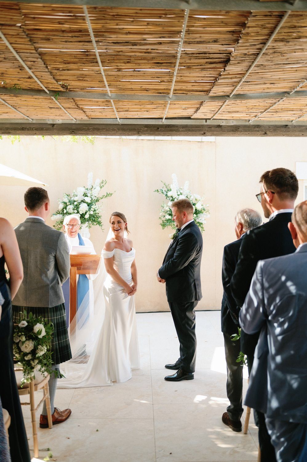 Wedding ceremony in progress under a rustic ceiling with white floral arrangements and standing guests.