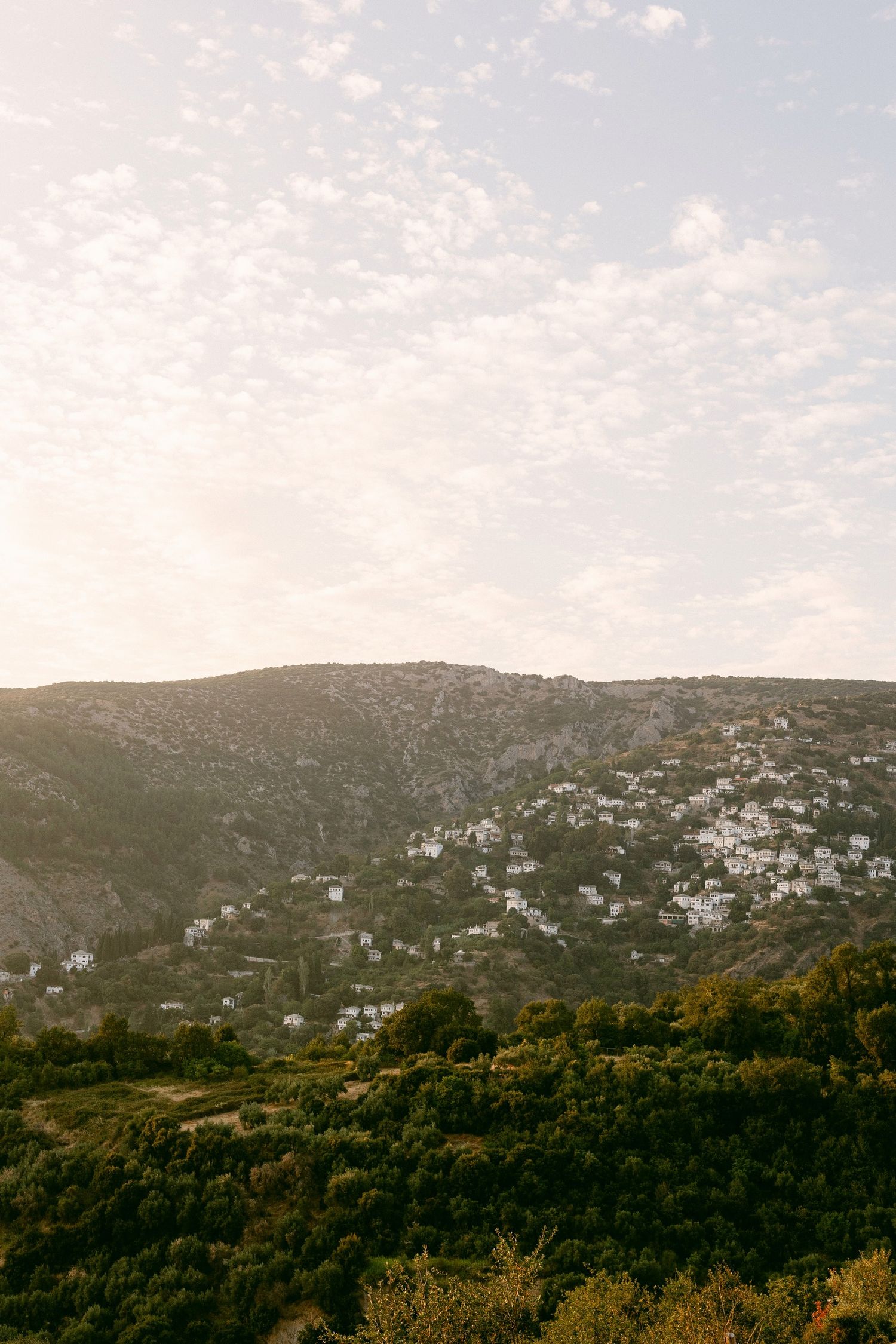 Houses dot the hillside of a scenic Mediterranean coastal town as golden sunlight illuminates the rugged terrain.