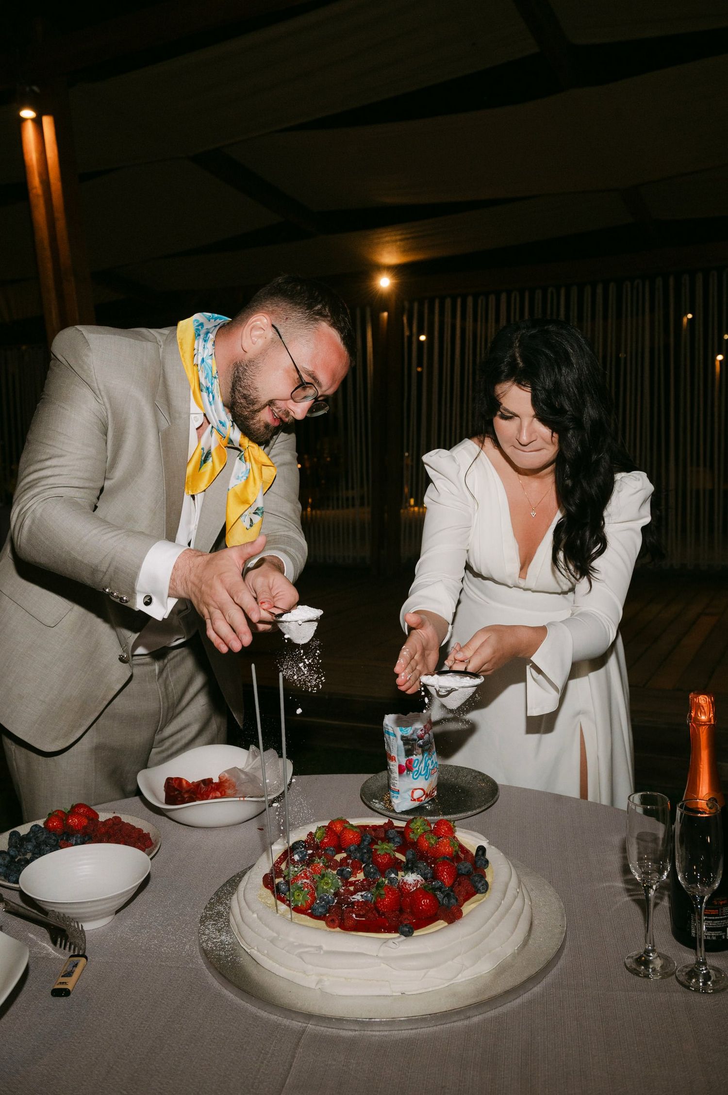 A couple cuts into a white pavlova dessert decorated with fresh strawberries and berries at an elegant dinner setting.