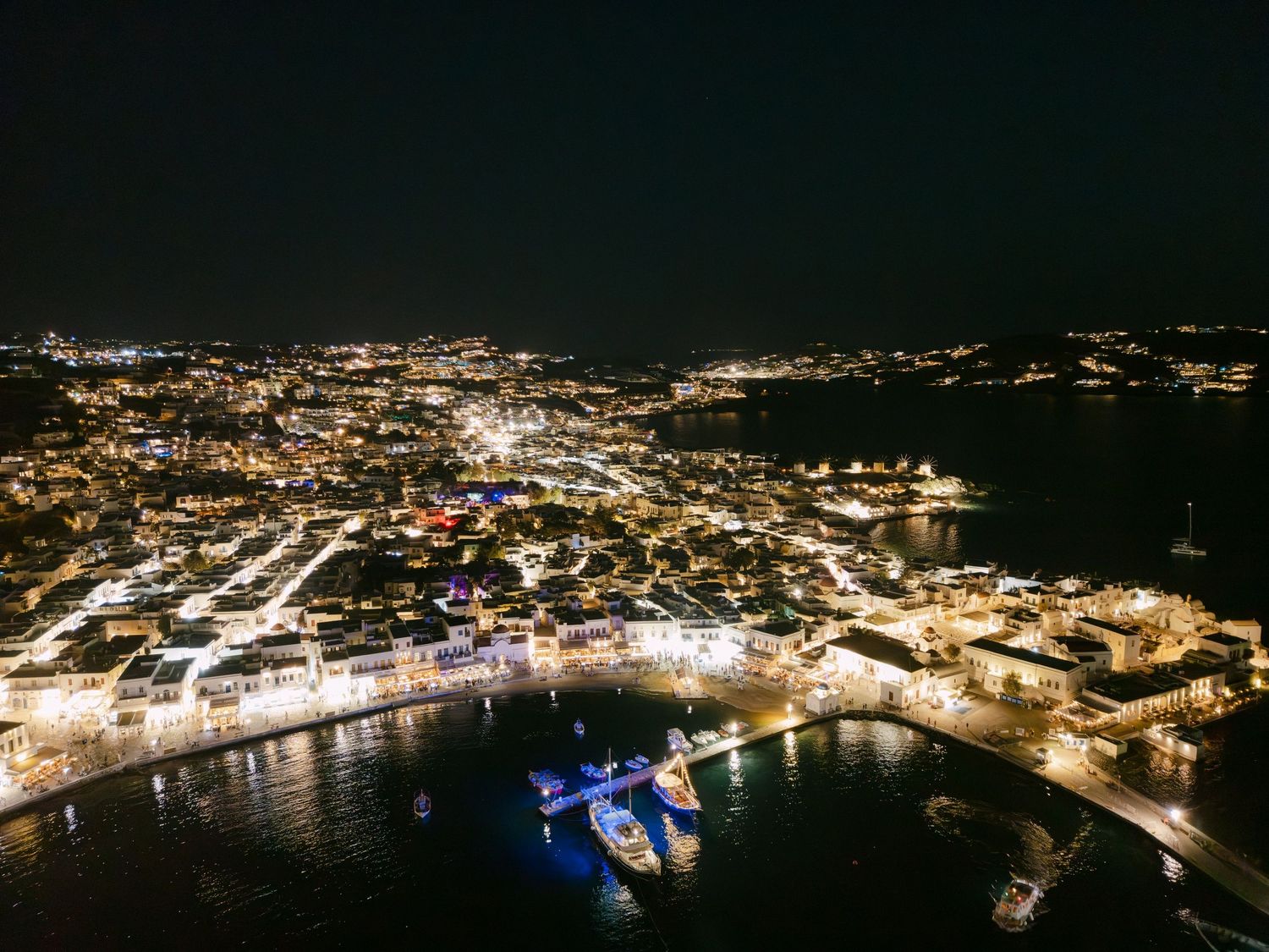 Aerial nighttime panorama of a coastal city with illuminated harbor and boats reflecting on calm waters.