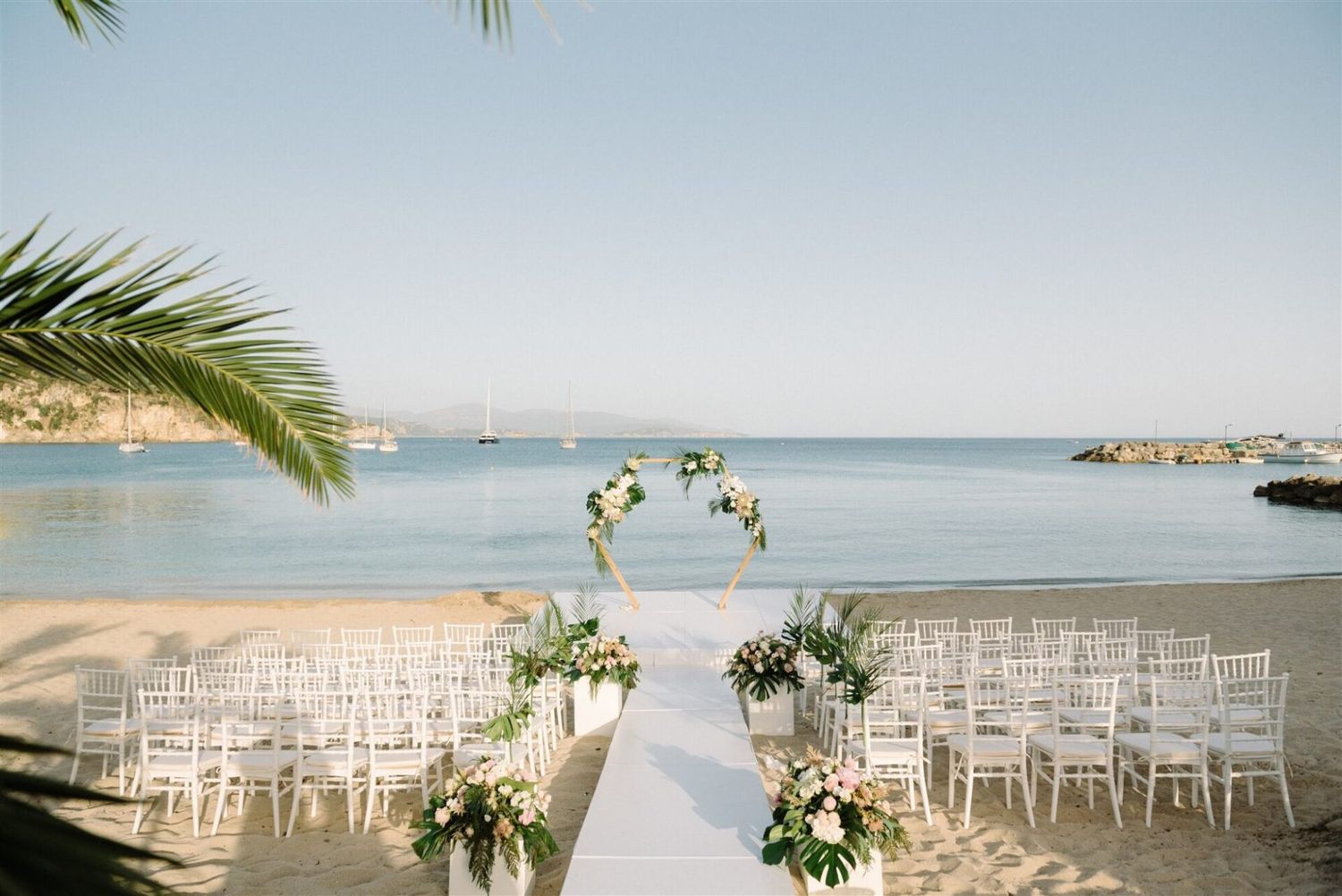 White chairs set up for a beach wedding ceremony with ocean views and palm trees framing the scene.
