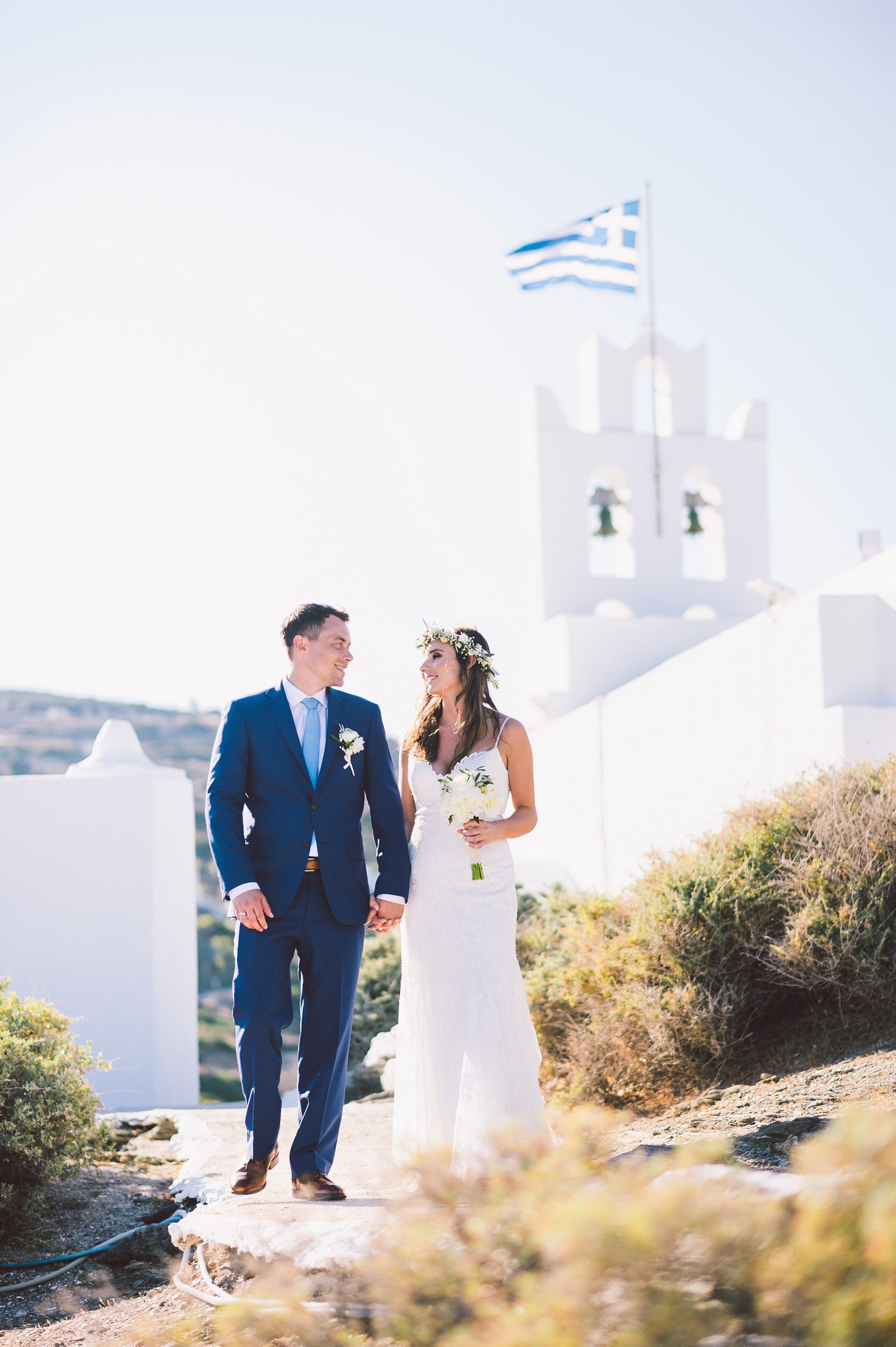 A couple in wedding attire walking together on a sunny hillside path at Chrysopigi chursch in Sifnos island in Greece.