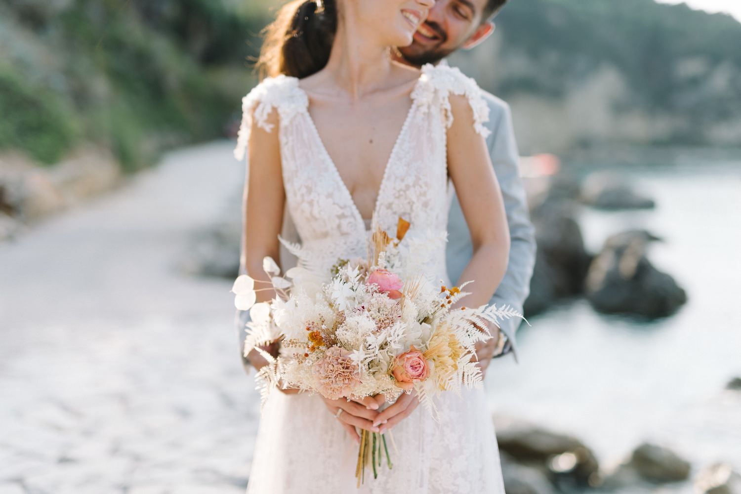 Bride in lace wedding gown holding a beautiful blush and cream floral bouquet on the beach.