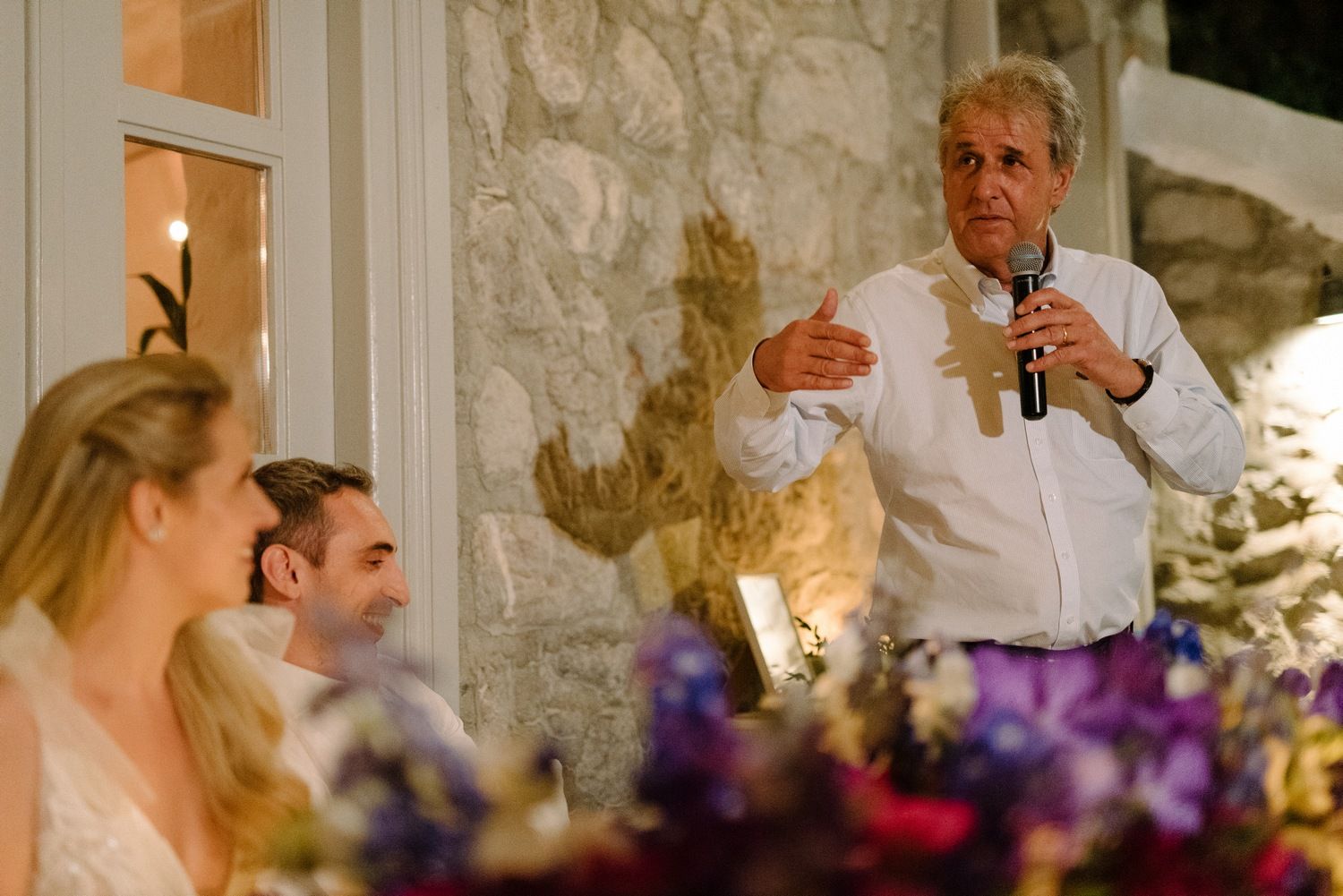 A toast being given during a wedding reception at a rustic stone venue with purple floral centerpieces.