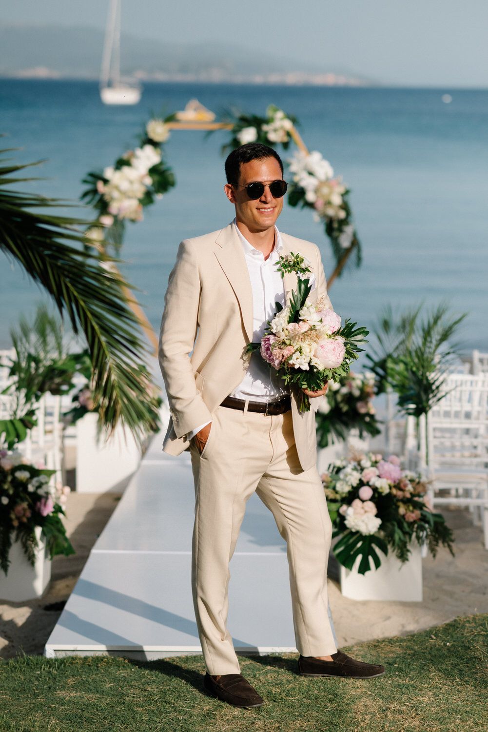 Person in beige suit holding bouquet stands by floral arch at beach wedding setup.