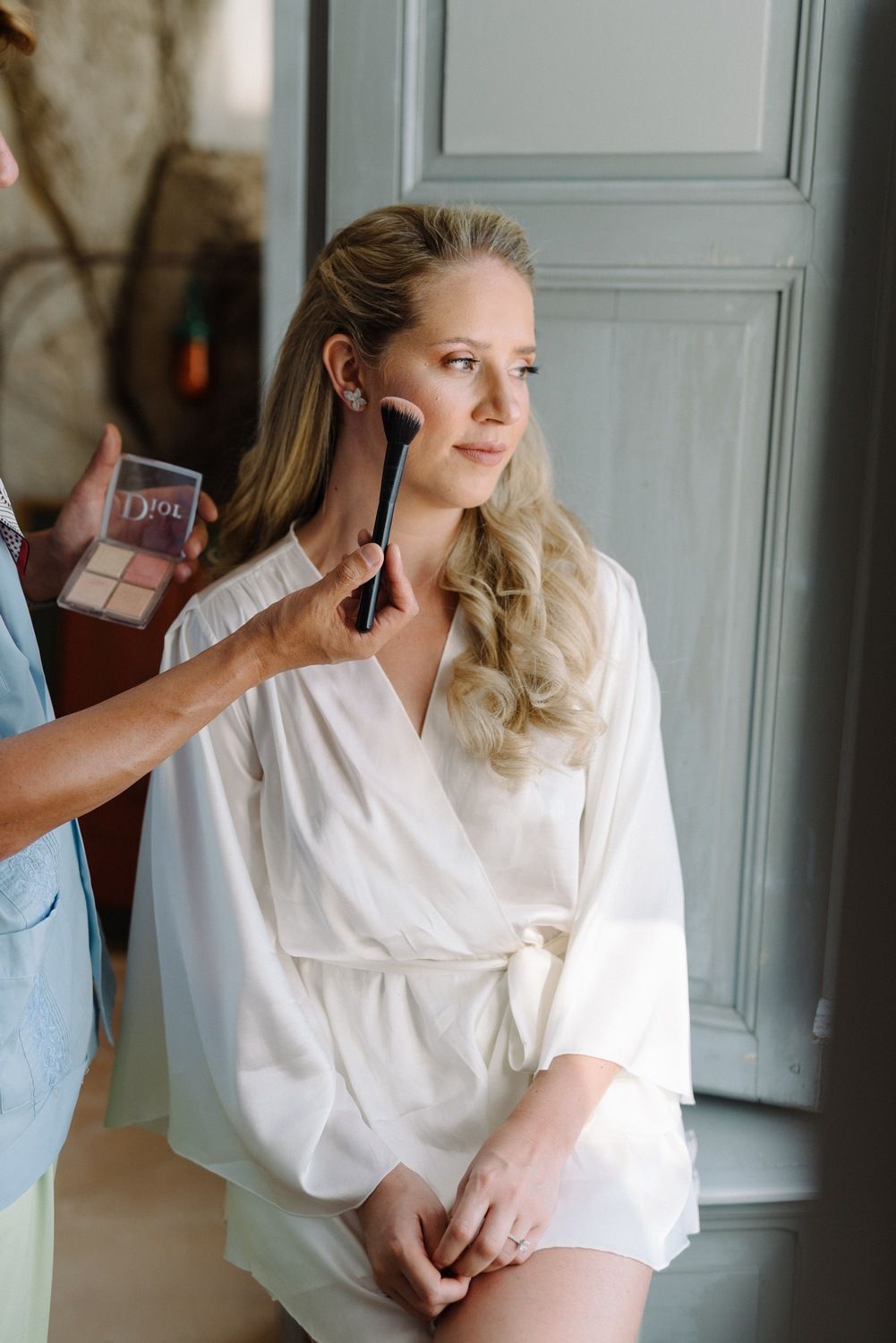 Makeup artist applying cosmetics on a woman in a white silk robe by a window.
